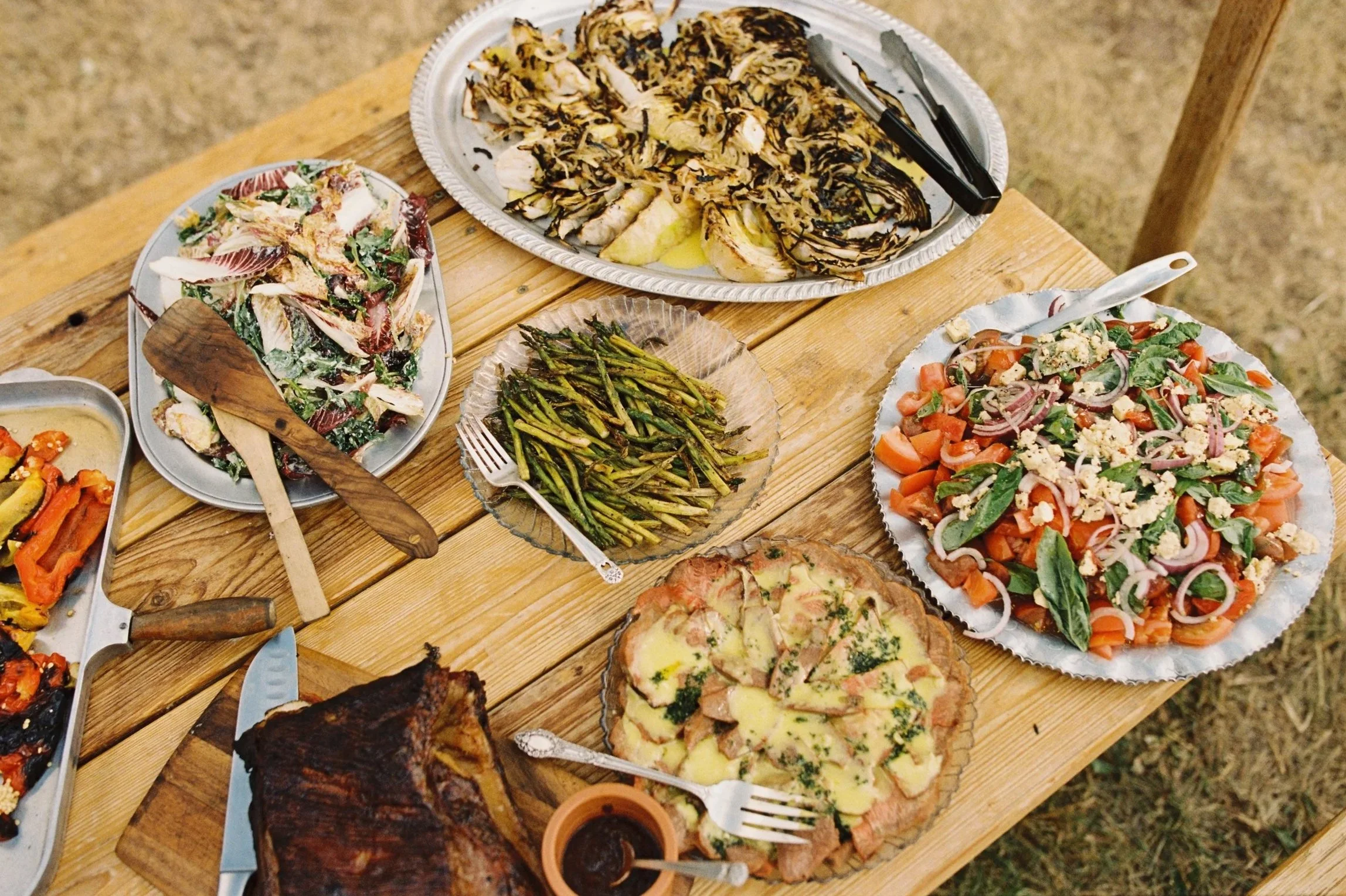 Assorted dishes including grilled vegetables, salad, roast beef, and other sides on a wooden table.