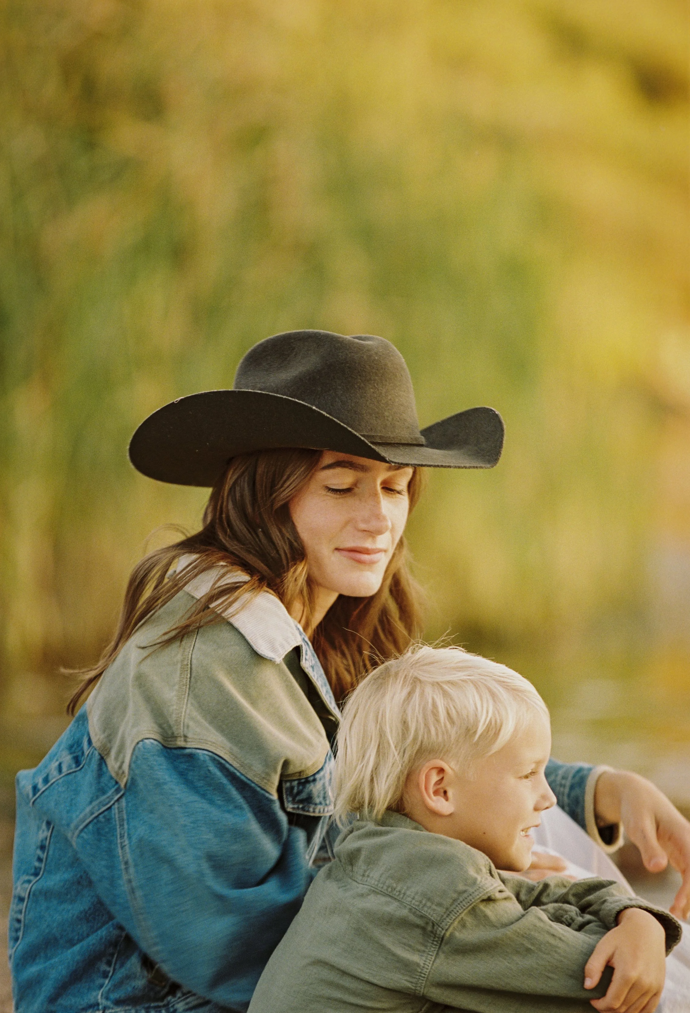 A woman wearing a large black hat and denim jacket sitting outdoors with a young boy who has blonde hair, both smiling and enjoying a moment together.