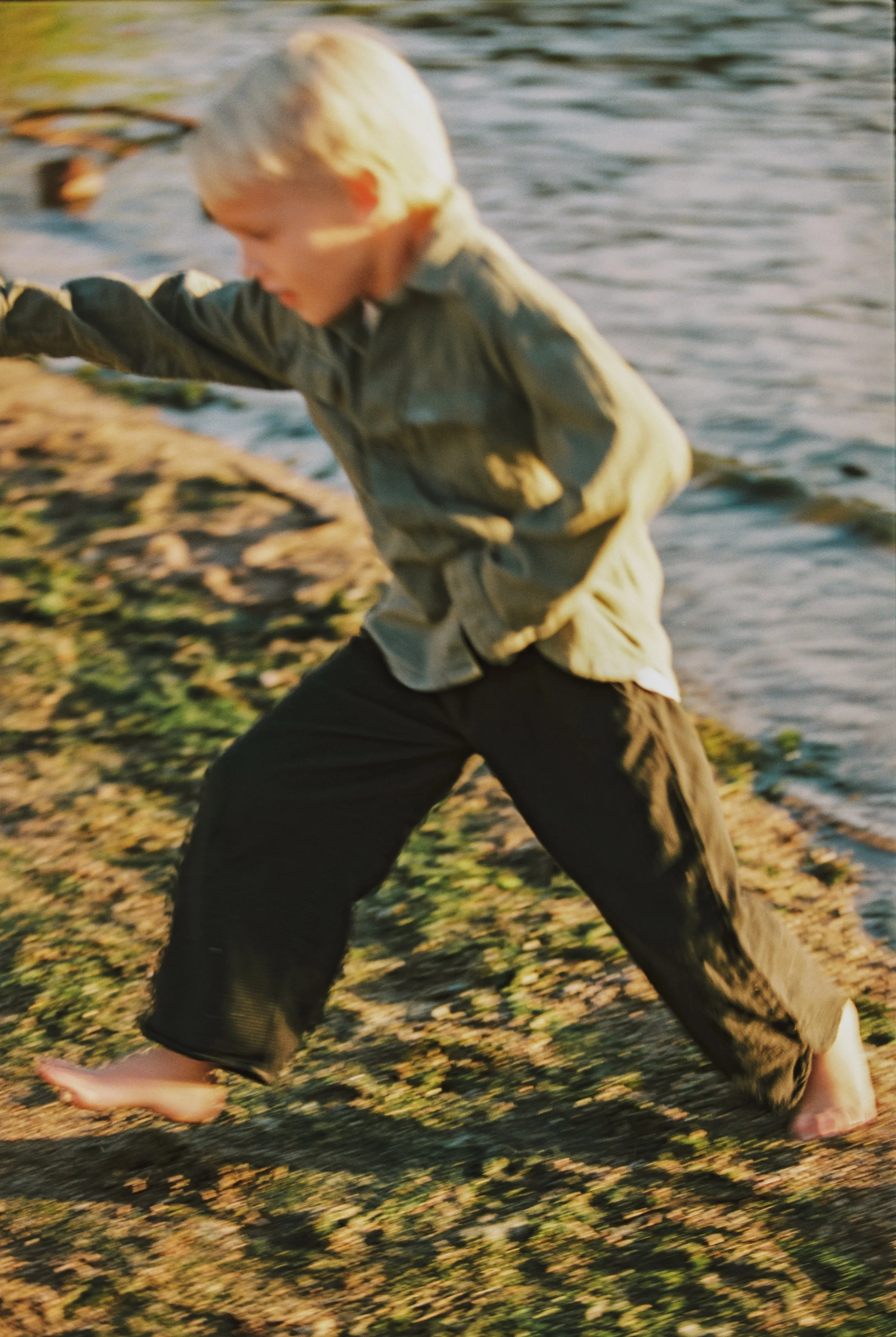 A young boy with blonde hair playing near the water's edge, barefoot, wearing a khaki shirt and black pants, stepping on wet sand and rocks.
