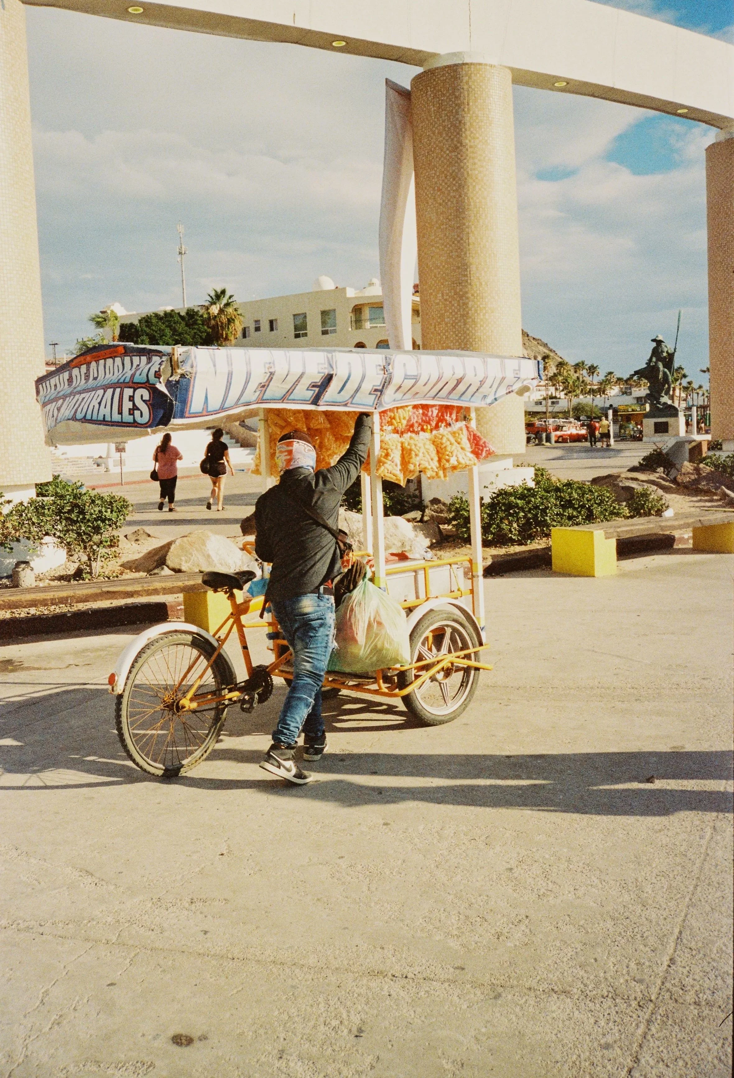 A person selling churros from a bicycle cart under a large covered structure in a public area, with a few pedestrians walking in the background.