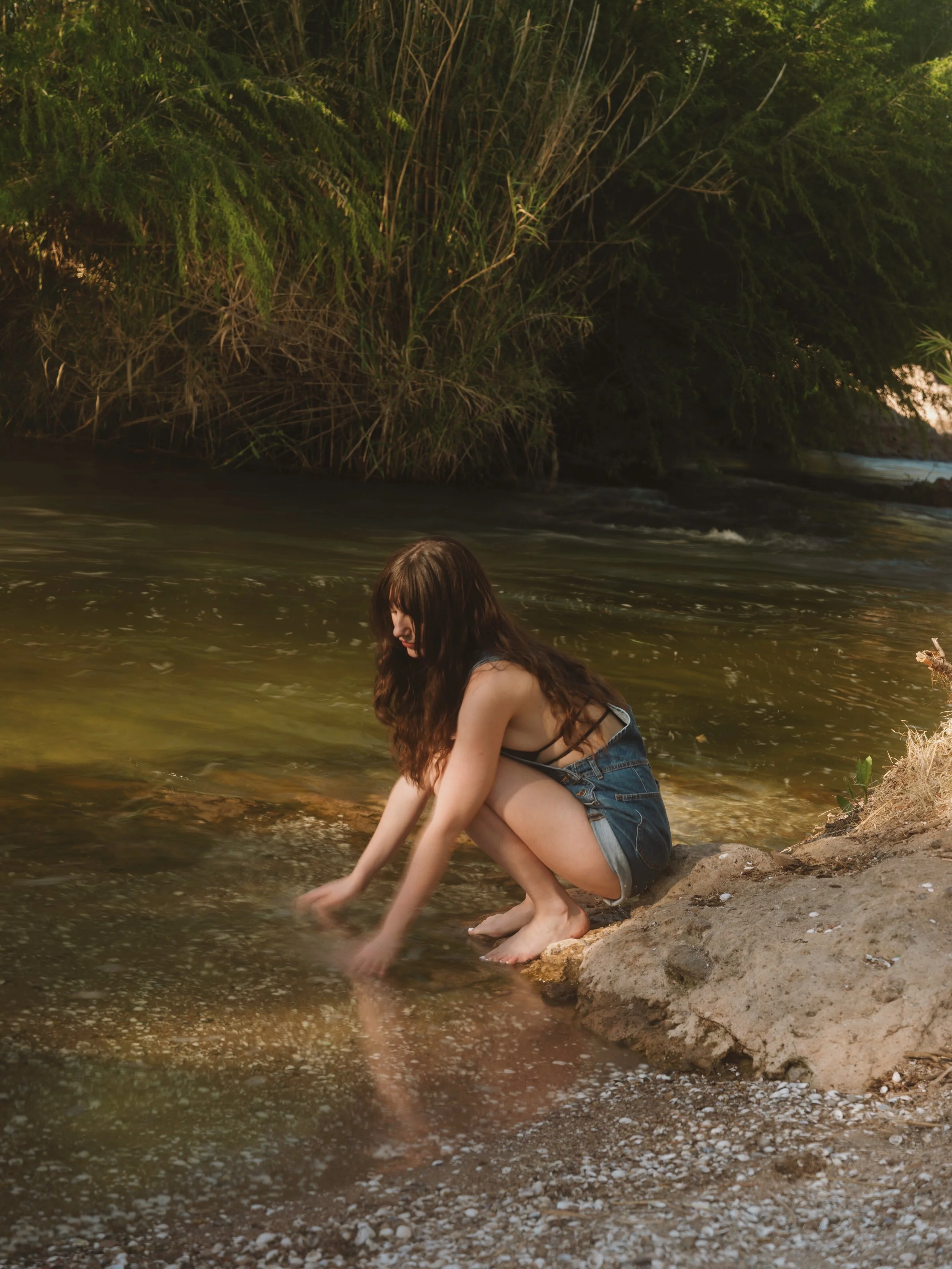 A young woman with long brown hair, wearing a black tank top and denim shorts, sitting on a rock at the edge of a river, reaching into the water during daytime with lush green trees in the background.