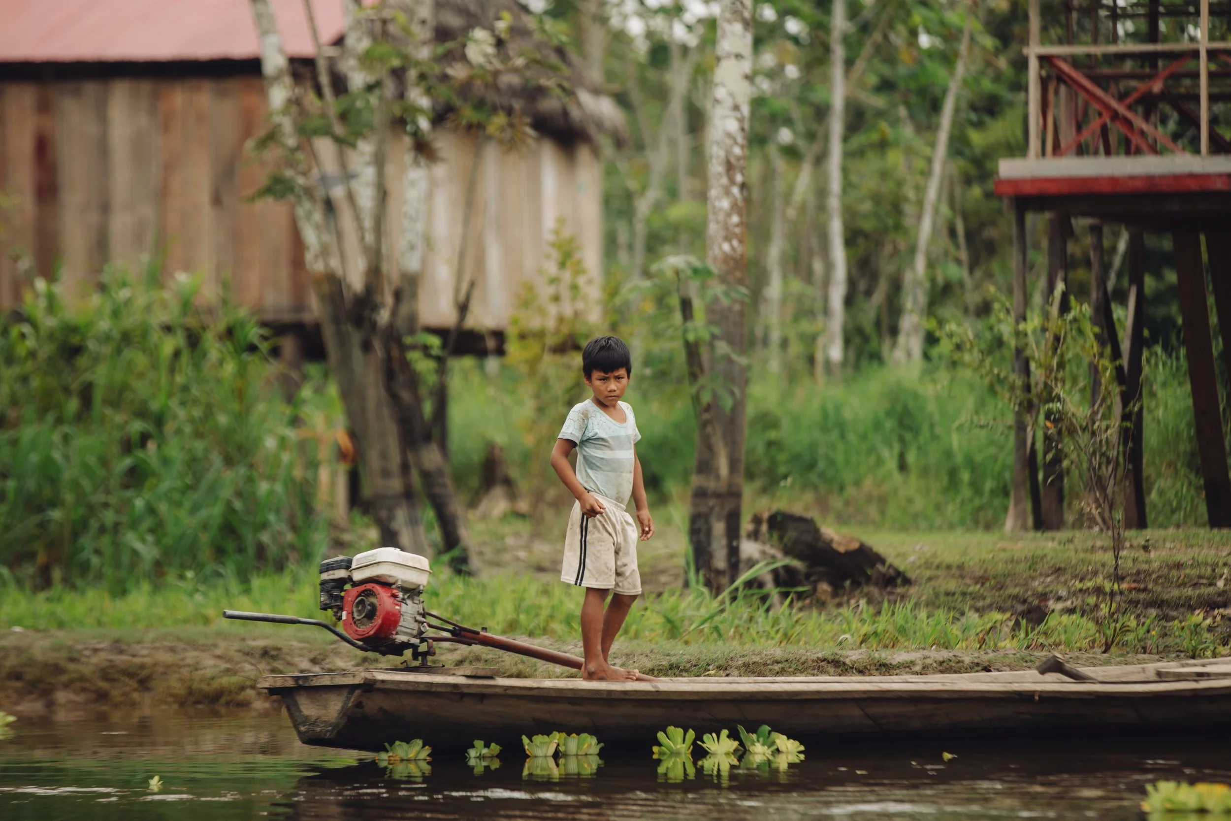 A young boy standing on a small wooden boat with a motor, on a canal or river, surrounded by lush greenery and wooden structures in the background.