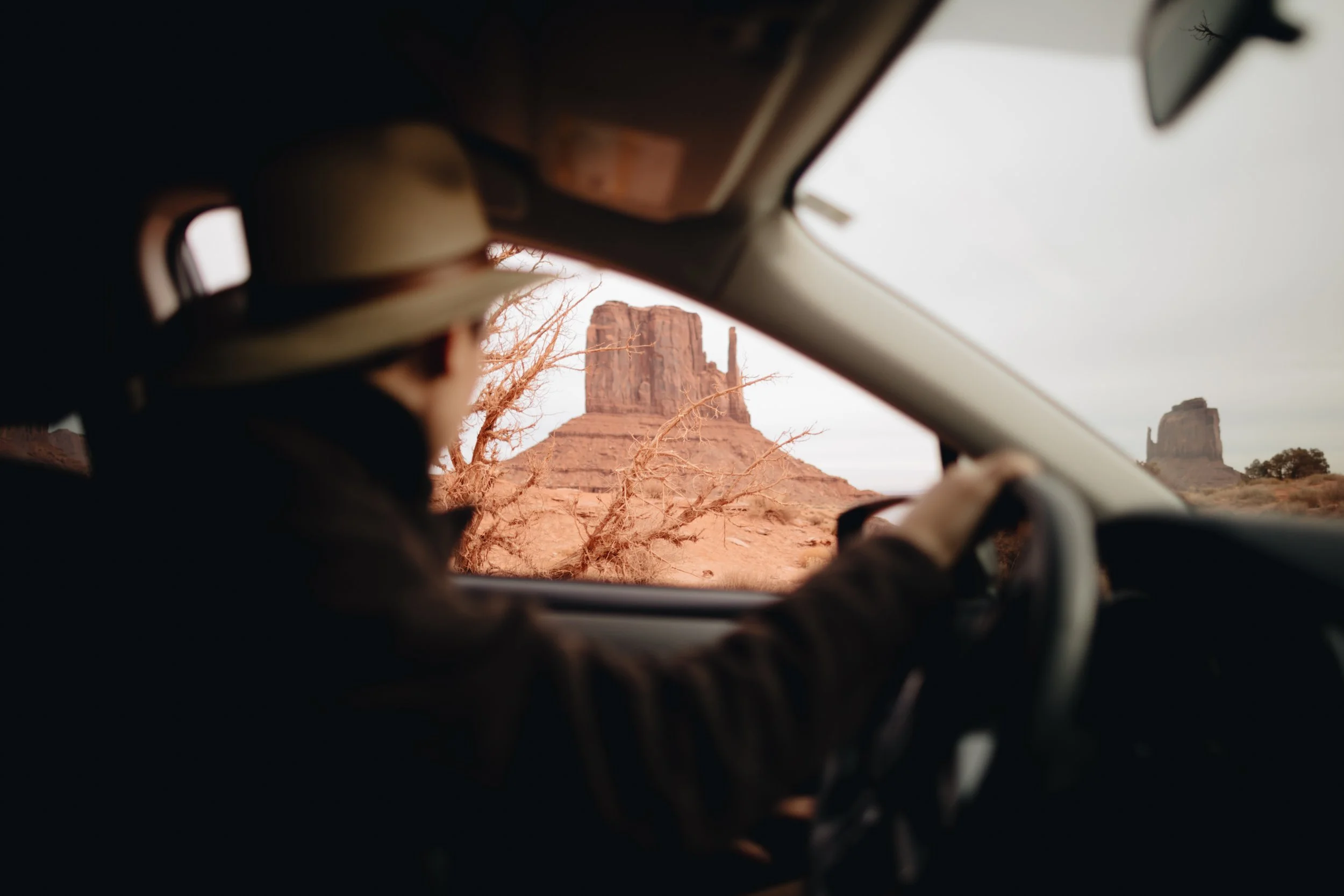 A person driving a car in a desert landscape with large rock formations, including a prominent butte, visible through the windshield.