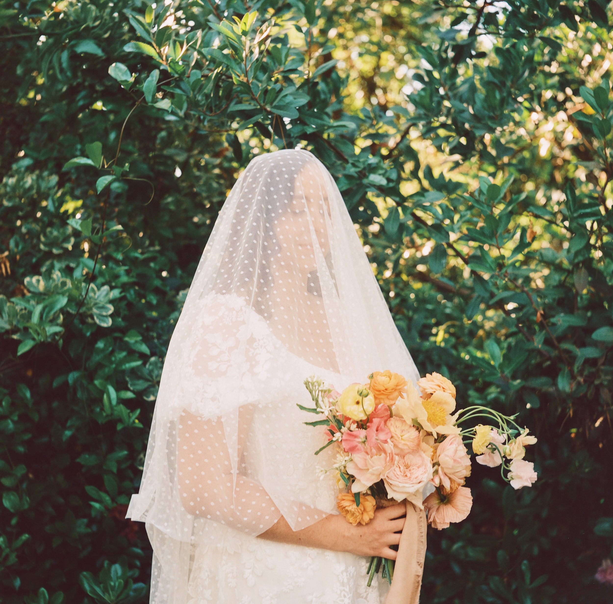 A bride in a wedding dress holding a bouquet of peach and pink flowers, standing outdoors with green foliage surrounding her, wearing a sheer polka dot veil that covers her face.