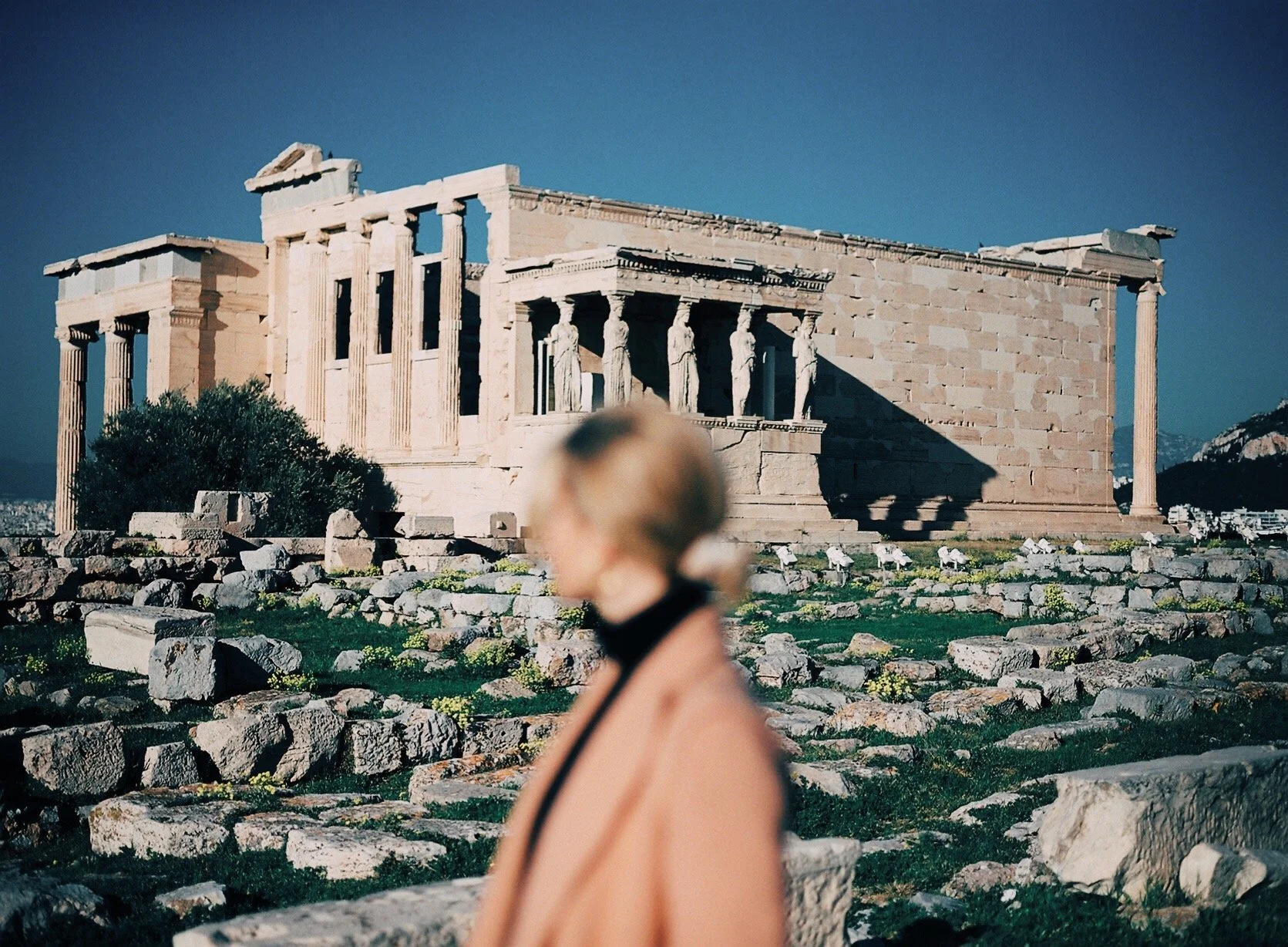 Ancient Greek temple ruins with multiple columns and sculptures, with a blurred woman in the foreground.