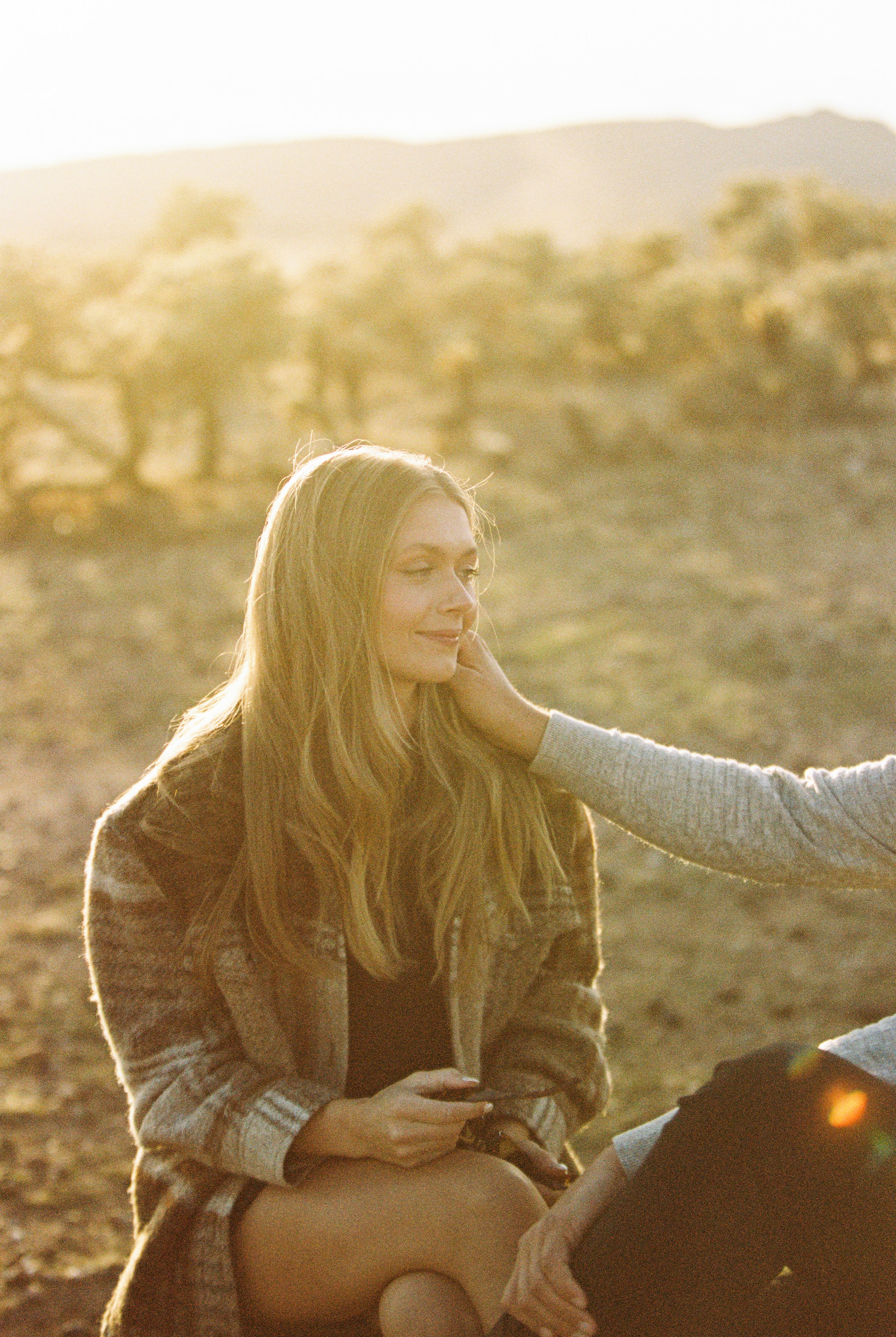 A woman with long blonde hair sitting outdoors on a sunny day, smiling gently as someone touches her face.