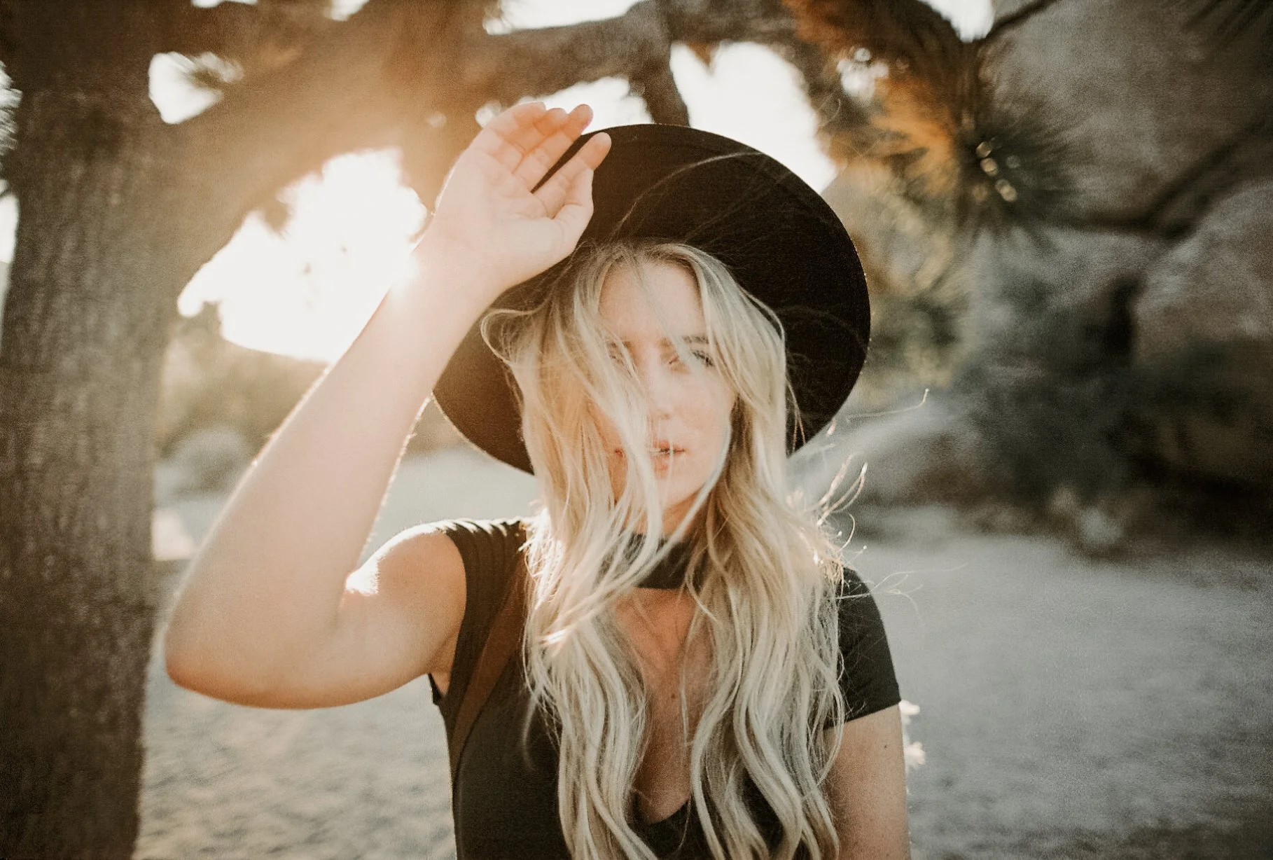 A woman with long blonde hair, wearing a black hat and black shirt, standing outdoors near a tree during sunset, with sunlight filtering through the branches.