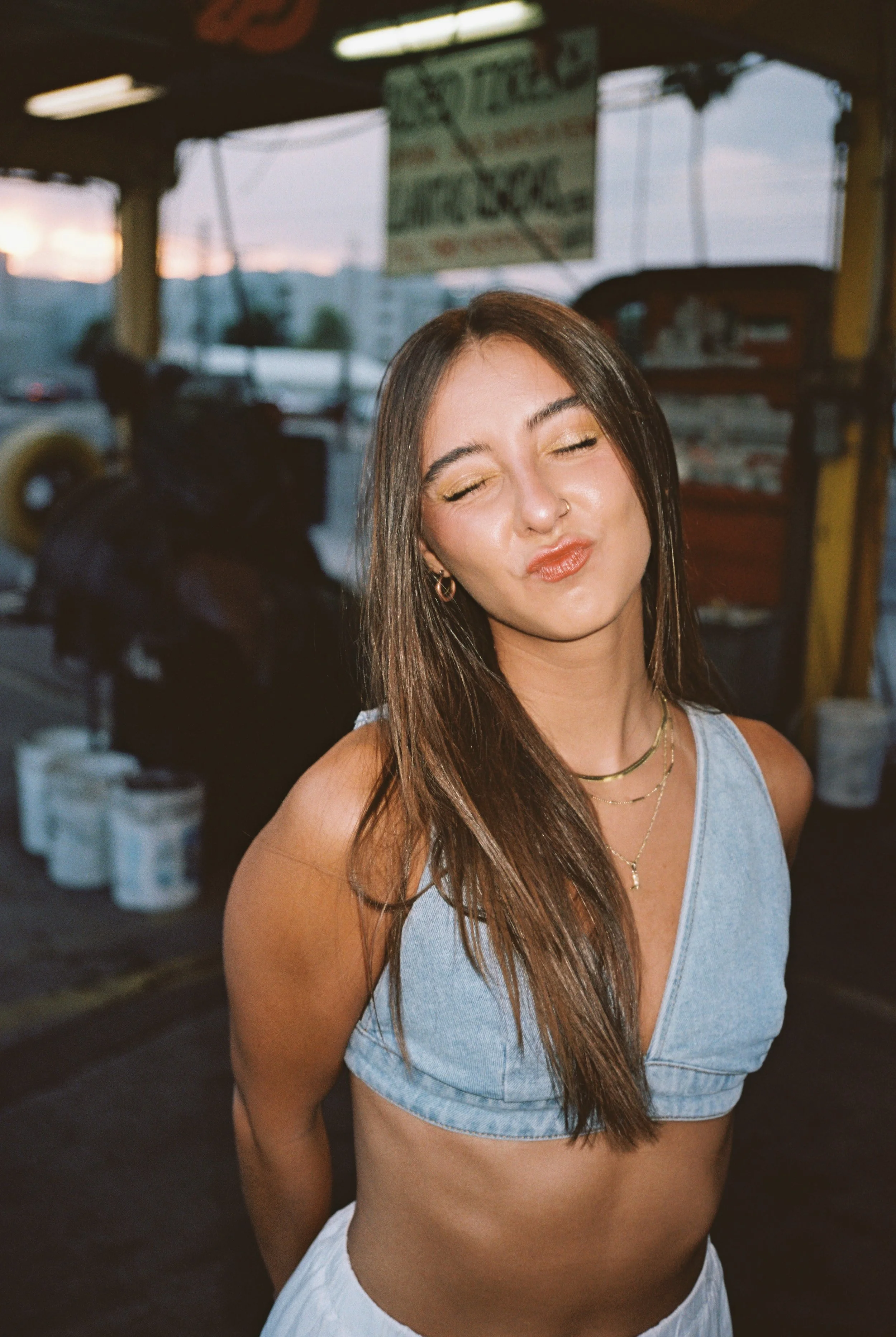 A young woman with long brown hair, closed eyes, and a playful pout, standing at a food stand or market during sunset, wearing a light blue crop top and layered gold necklaces.