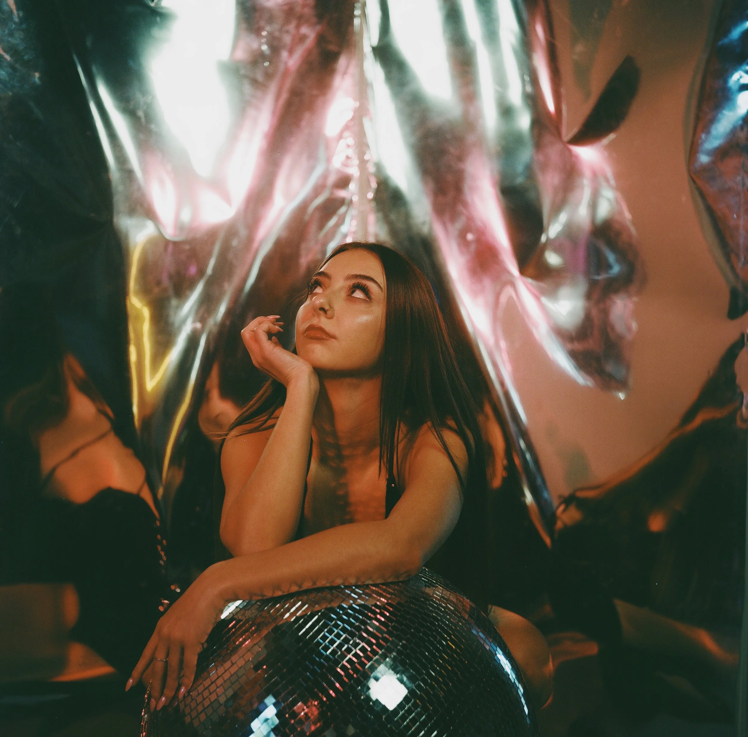 A young woman with long dark hair sitting among metallic, reflective surfaces with a disco ball in front of her, looking upward thoughtfully.