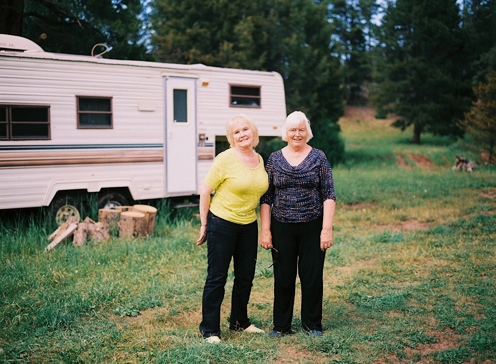 Two elderly women standing outdoors in front of a white camper trailer, smiling and posing for the photo with trees and grass in the background.