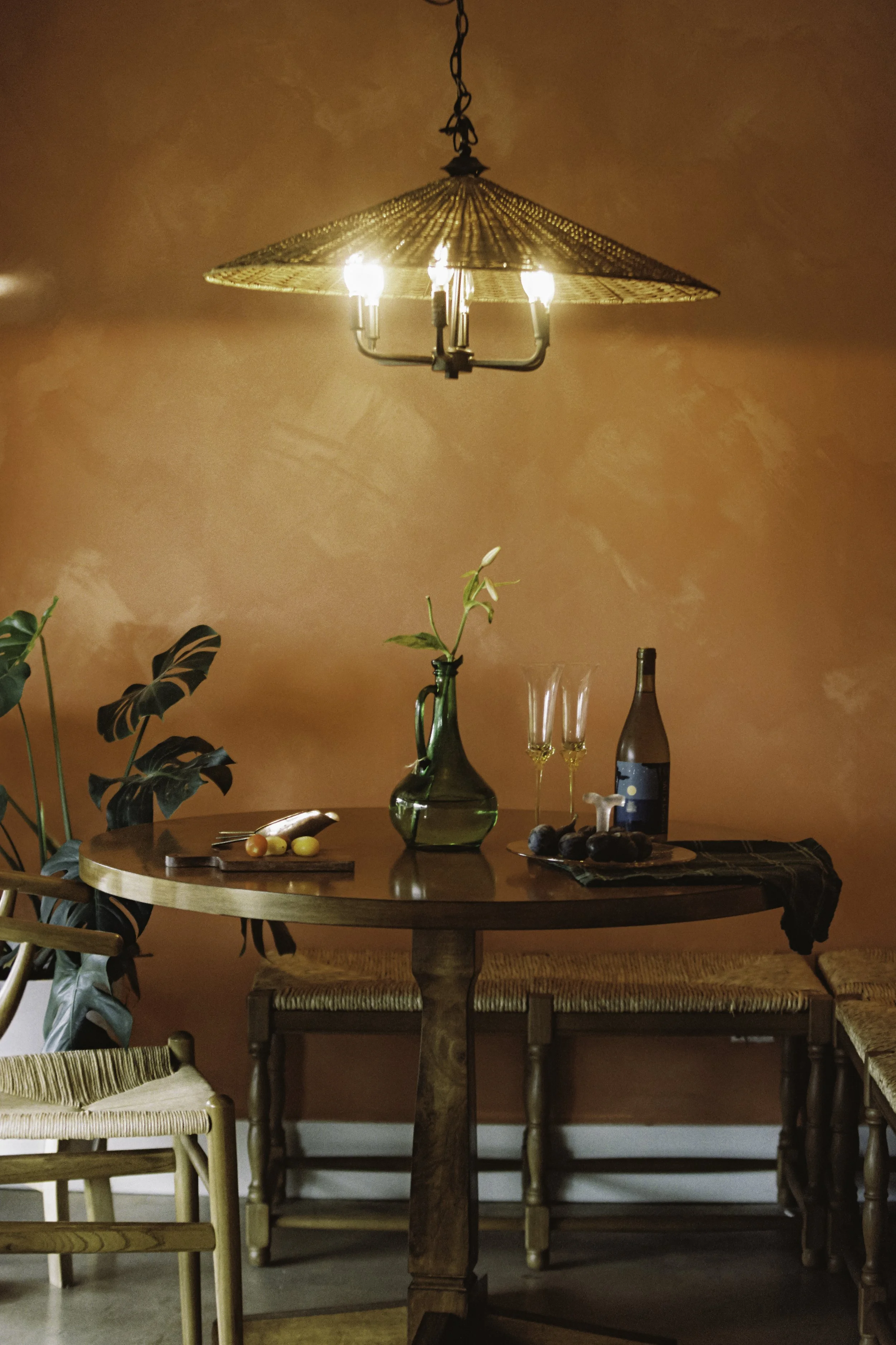 A wooden dining table with a green vase holding white flowers, two champagne glasses, a wine bottle, and a plate of figs. A rattan lampshade chandelier hangs above the table, and there are wooden chairs around the table against an orange textured wal