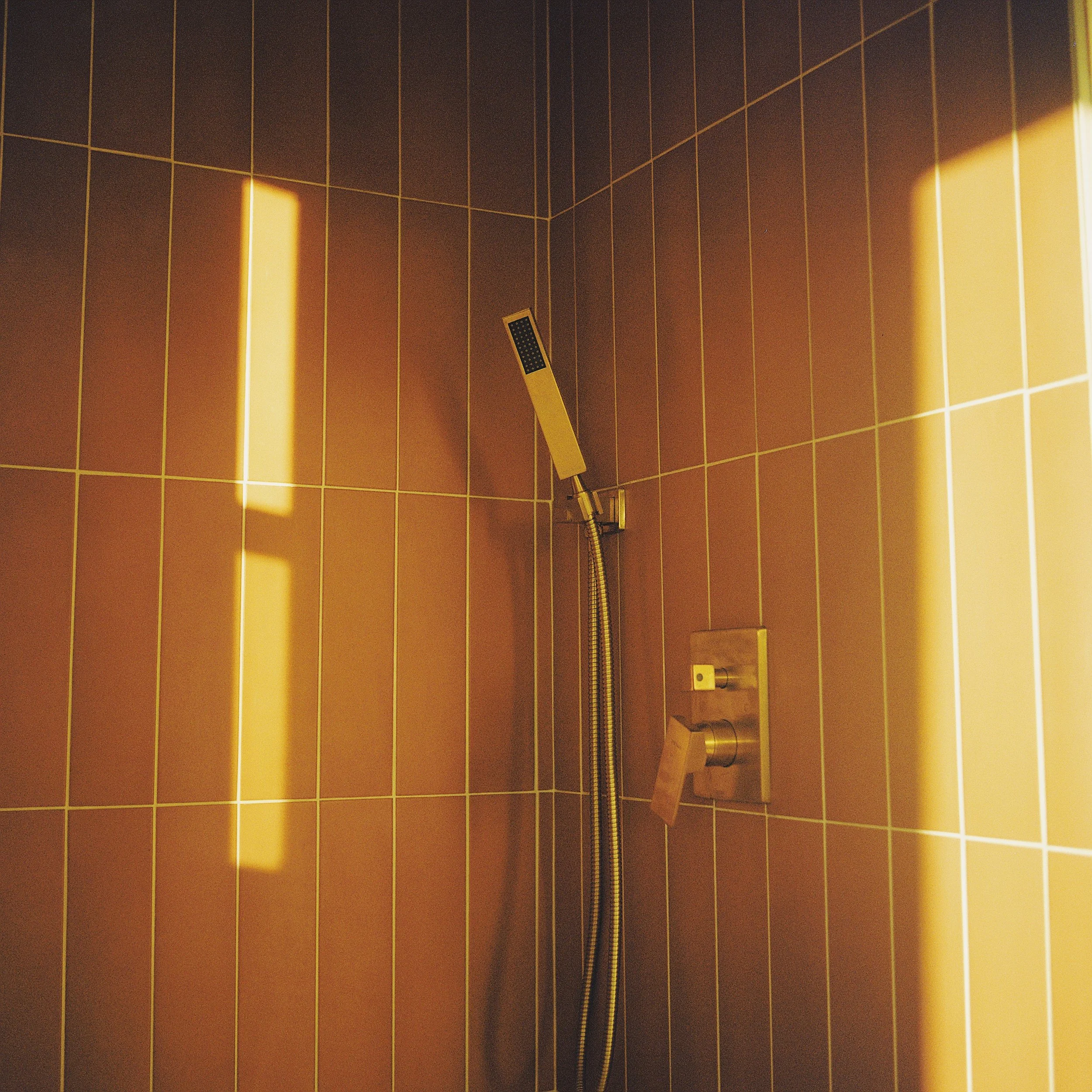 Shower with brown tiled walls, a yellow handheld showerhead, and sunlight casting shadows on the tiles.