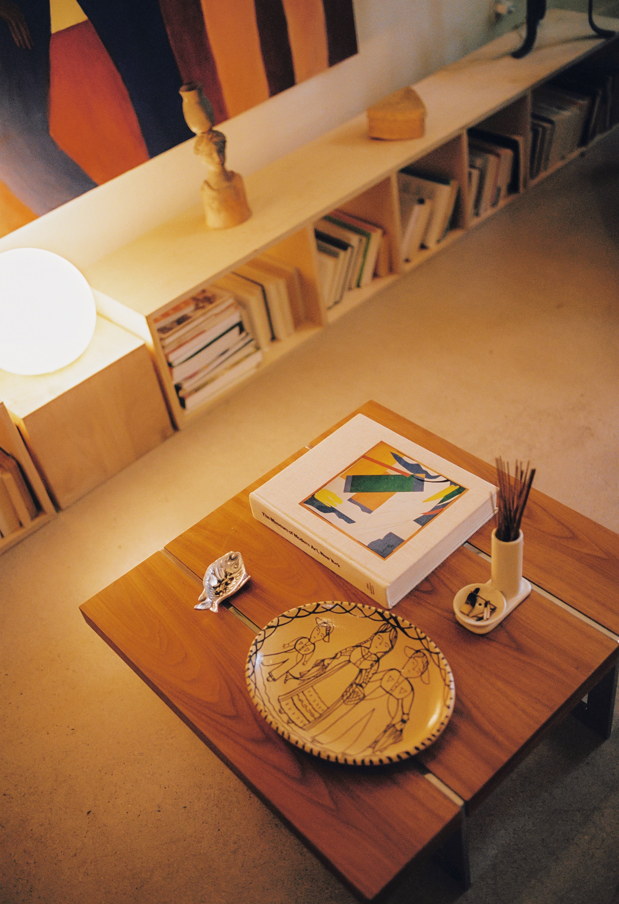 A wooden table with a colorful book, a yellow decorative plate with black line art, a small white vase holding incense sticks, and a metallic fish figurine. In the background, a wooden bookshelf with books and a spherical lamp, with part of a abstrac