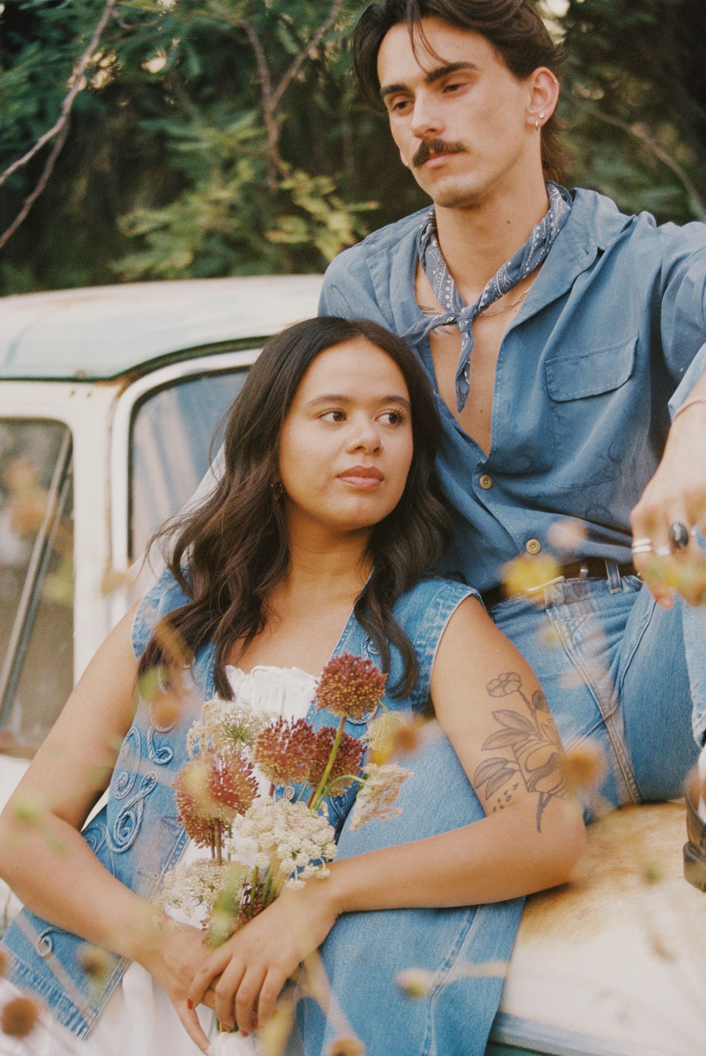 A woman with long dark hair, tattoos on her arm, wearing a denim vest, holding a bouquet of flowers, sitting in front of a vintage car. A man with a mustache, earrings, and a blue shirt with a bandana around his neck stands beside her, resting his ar