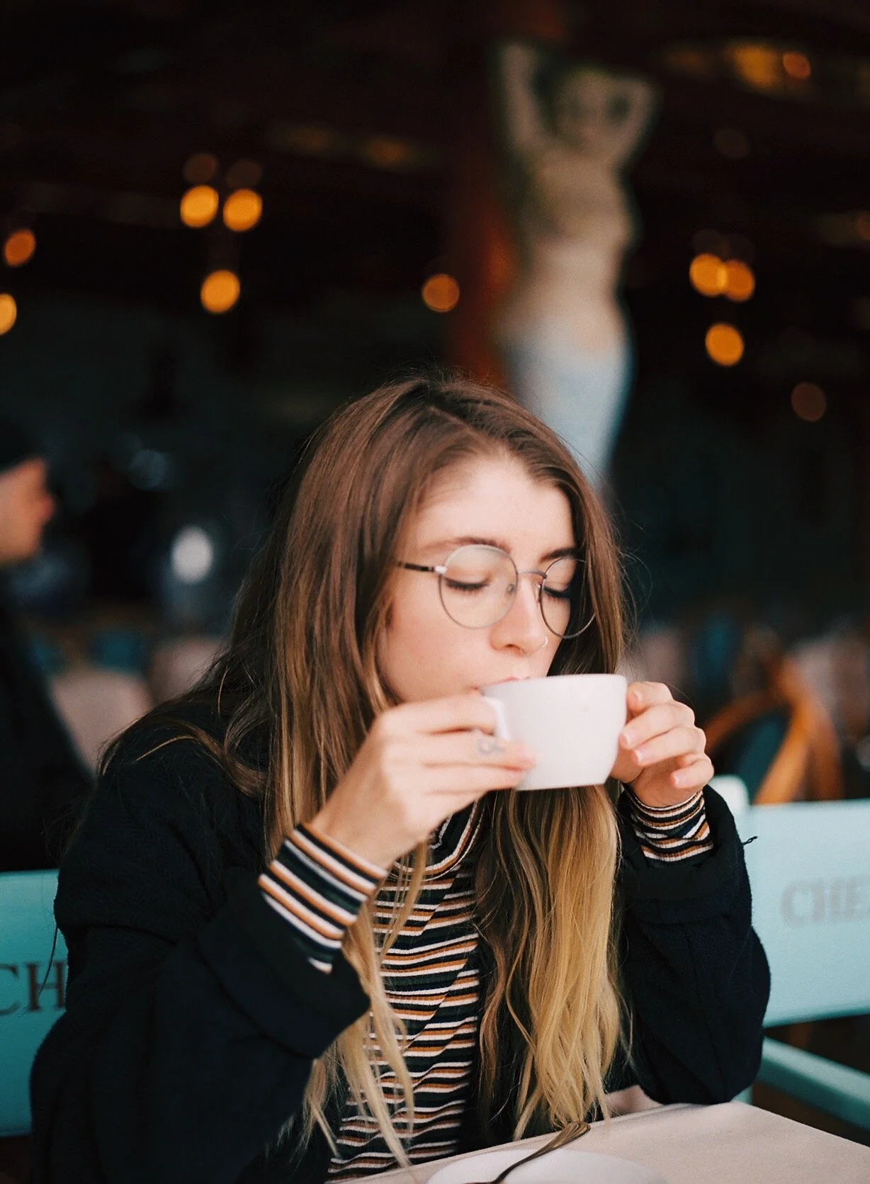 A young woman with long blonde hair, wearing glasses and a striped shirt, drinking from a white mug in a cozy cafe.