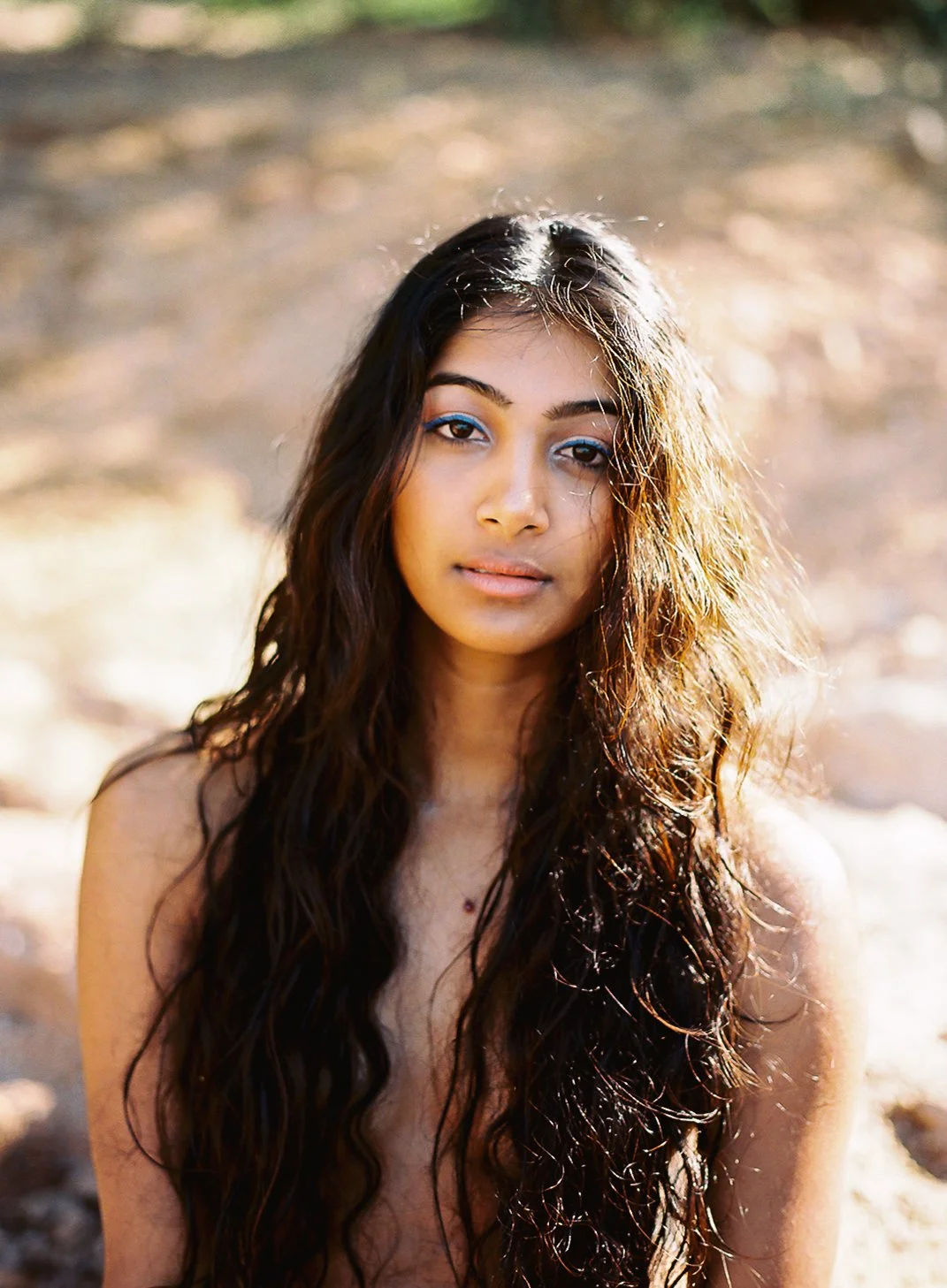 A young woman with long, wavy dark hair outdoors, looking at the camera with a neutral expression. The background is blurred with natural sunlight.