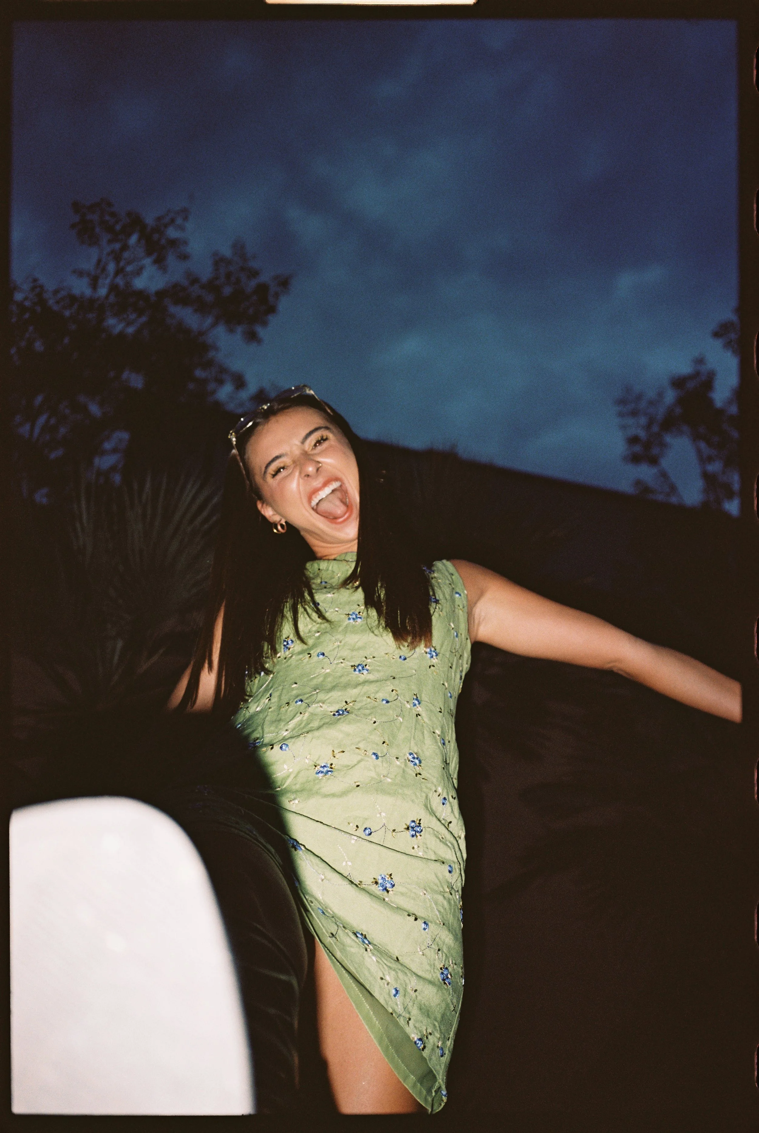 A woman in a green floral dress celebrating outdoors at night, smiling and making a playful face with her tongue out, with a dark sky and trees in the background.