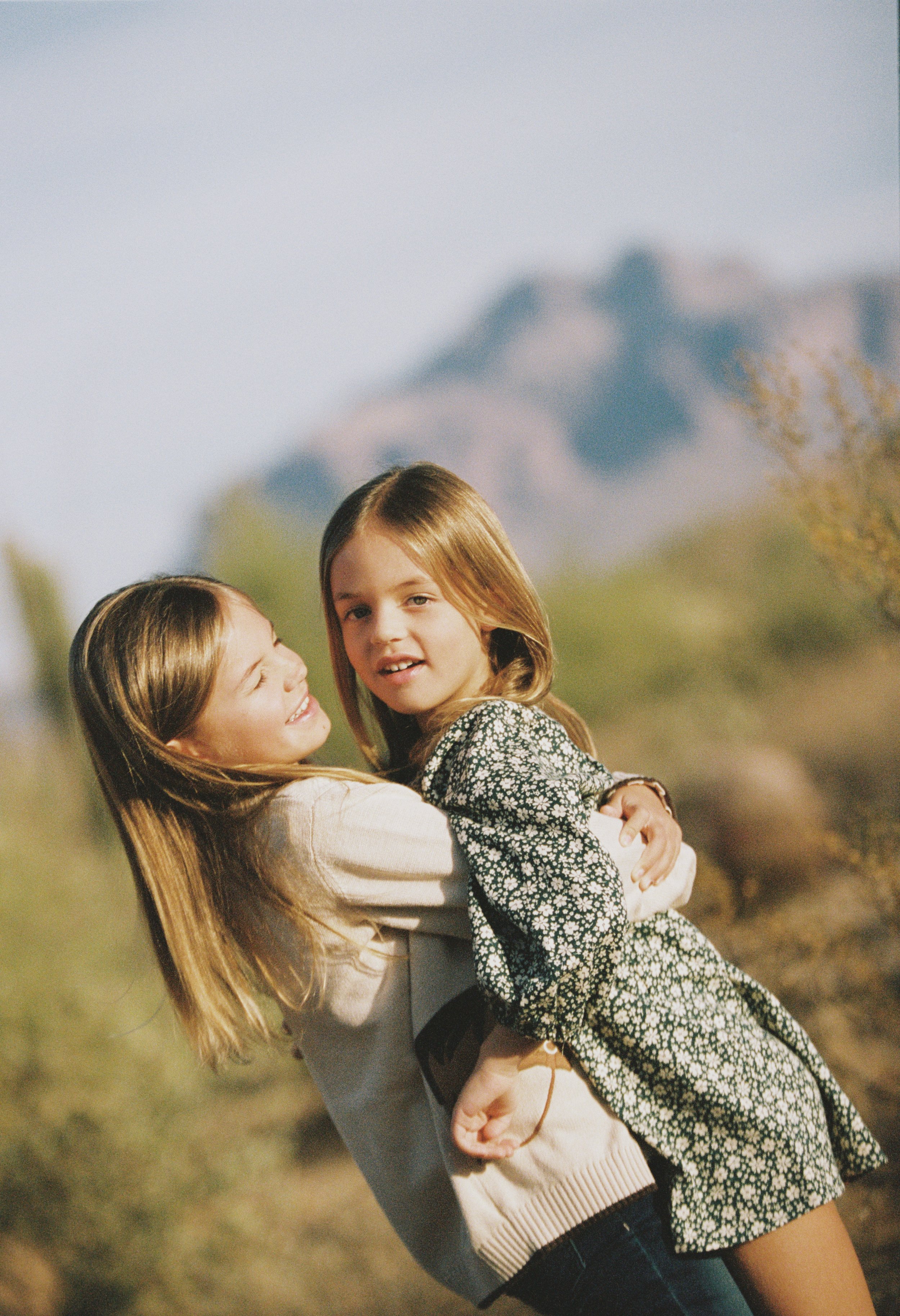 Two young girls outdoors, one holding the other in her arms, with a mountain in the background.