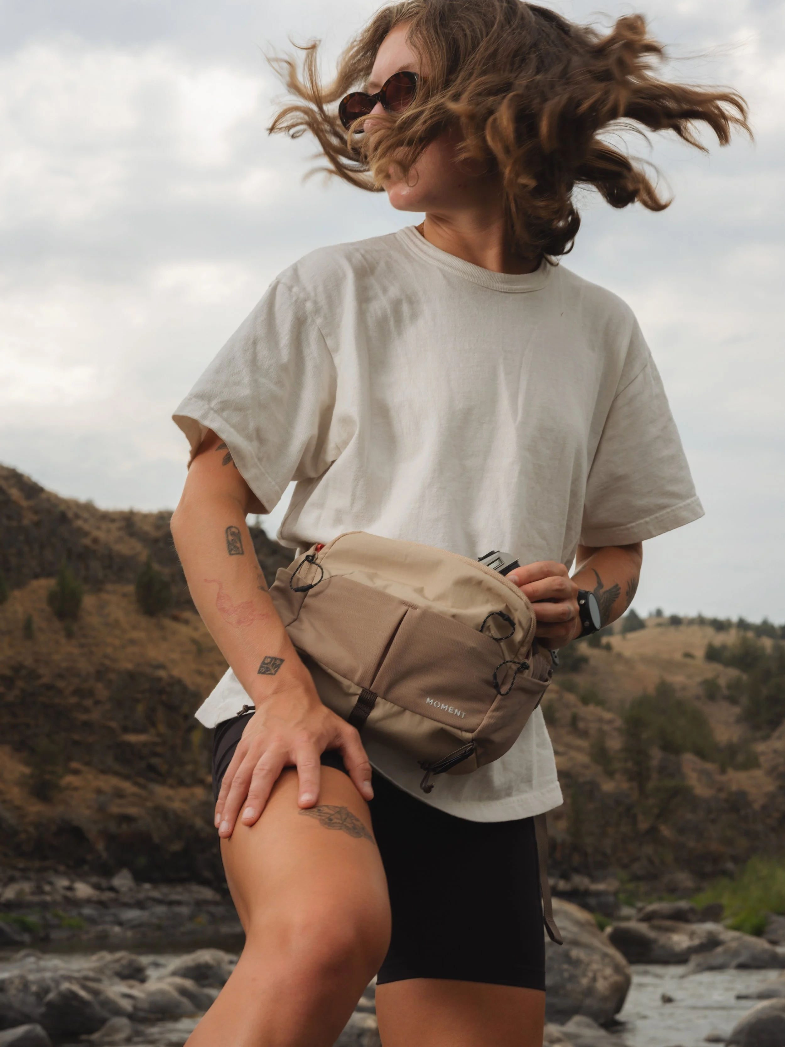 Woman with curly hair wearing sunglasses, a loose white t-shirt, and black shorts, holding a beige backpack outdoors near a river and rocky landscape.