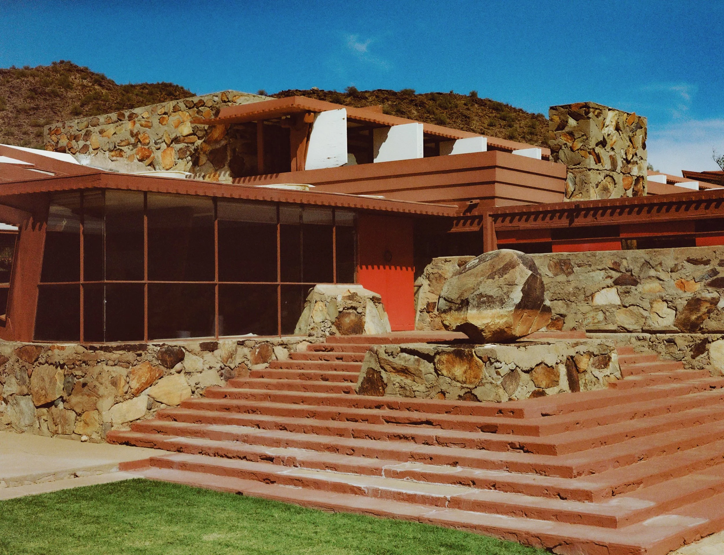 A modern house built with stone and red stucco, featuring large windows, set against a rocky hillside and clear blue sky.