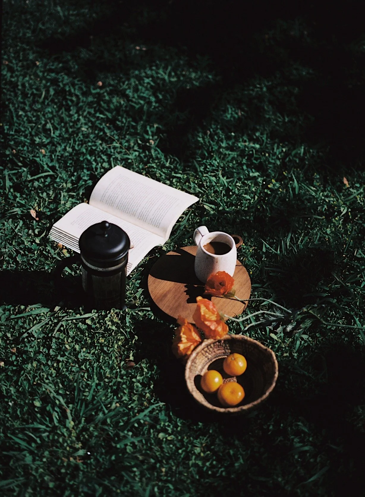 A picnic setup on grass with an open book, a black thermos, a white mug of coffee, a small wooden tray, an orange flower, a basket of yellow fruit, and some orange flowers.