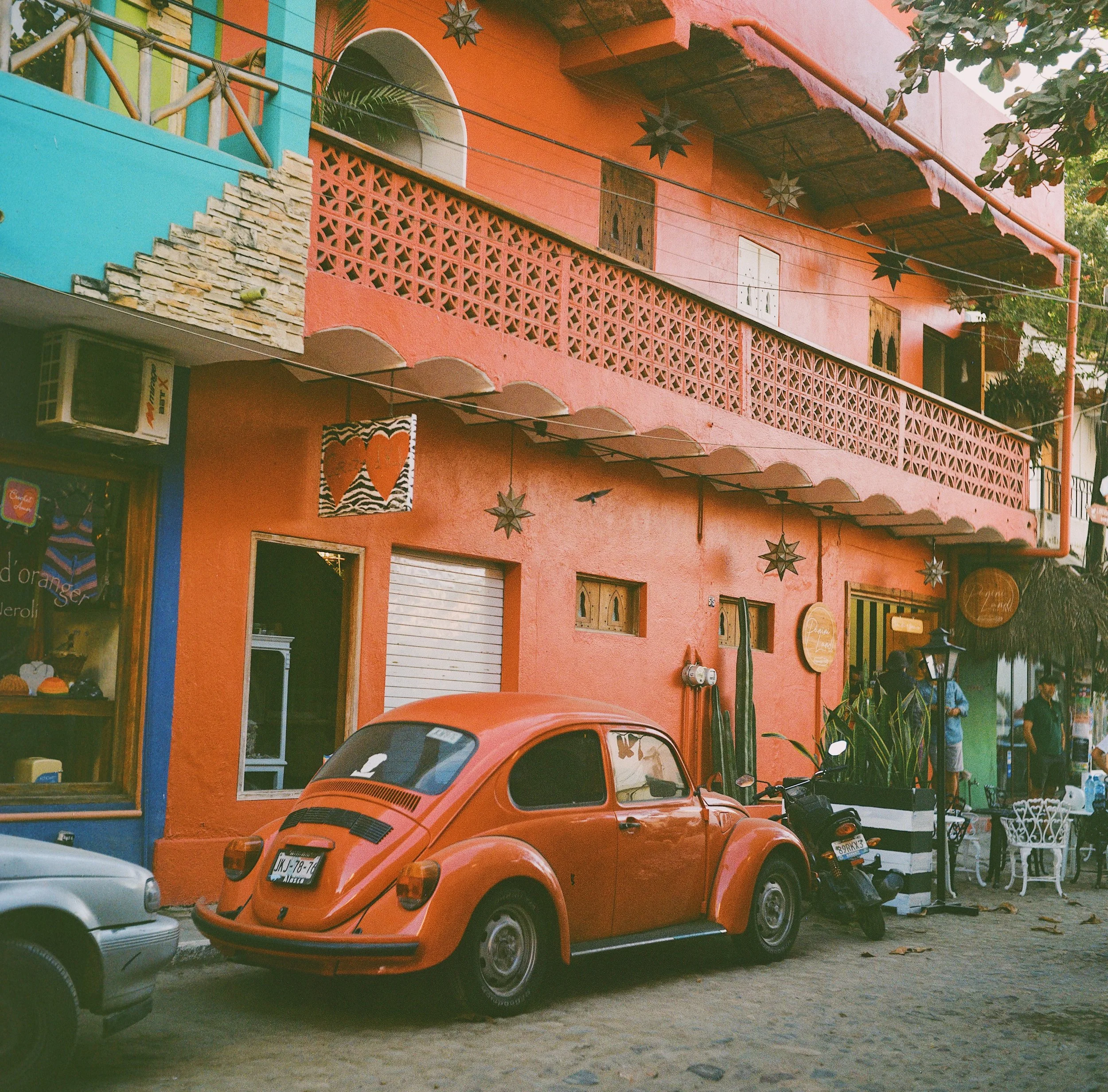 A vintage orange Volkswagen Beetle parked on a street in front of a colorful building with pink and blue walls, decorative stars hanging from the balcony, and people walking nearby.