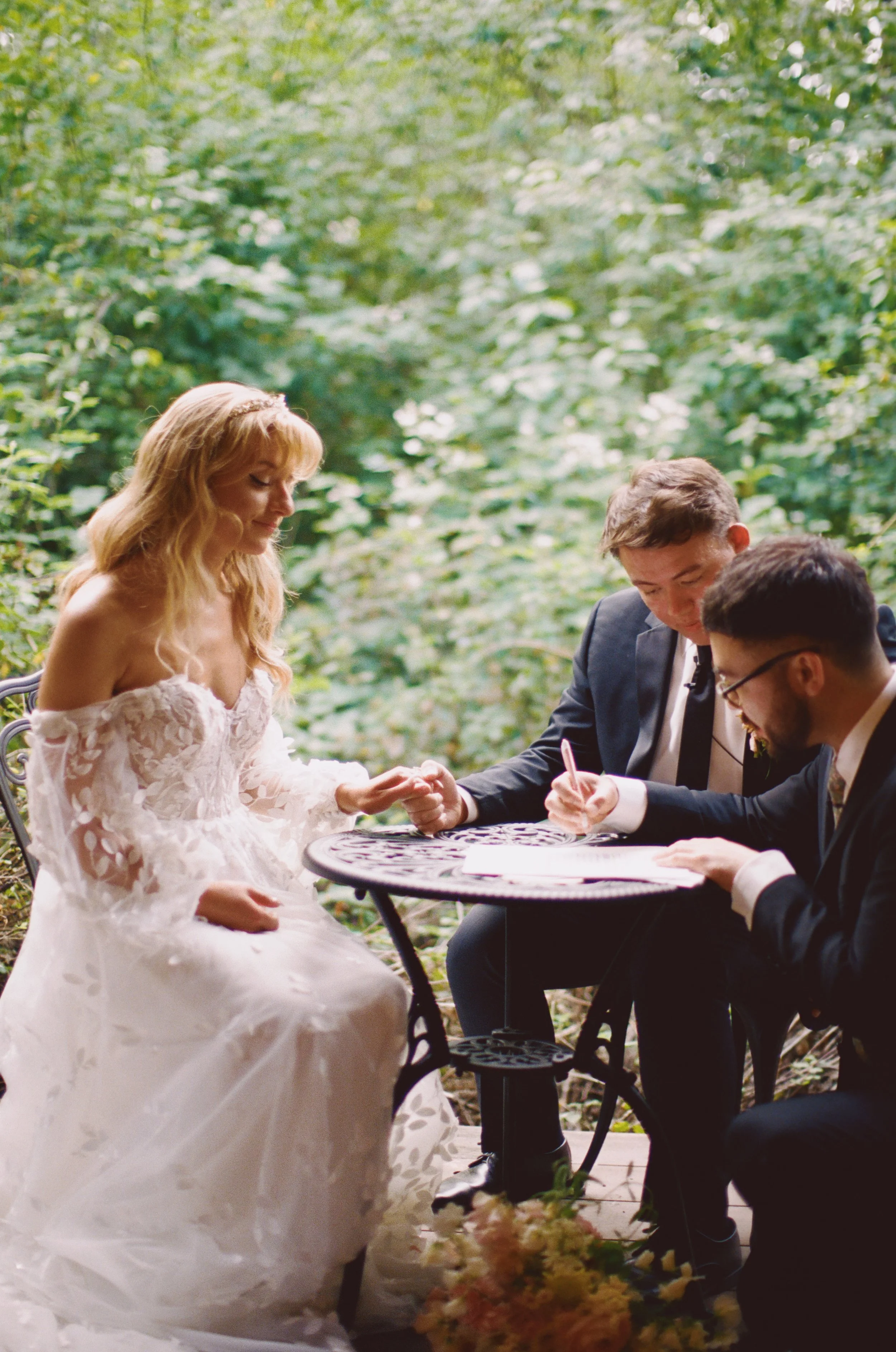 A wedding ceremony outdoors with a bride in a white lace dress, sitting at a small round table with two men in suits, one of whom is signing a document, surrounded by green foliage.