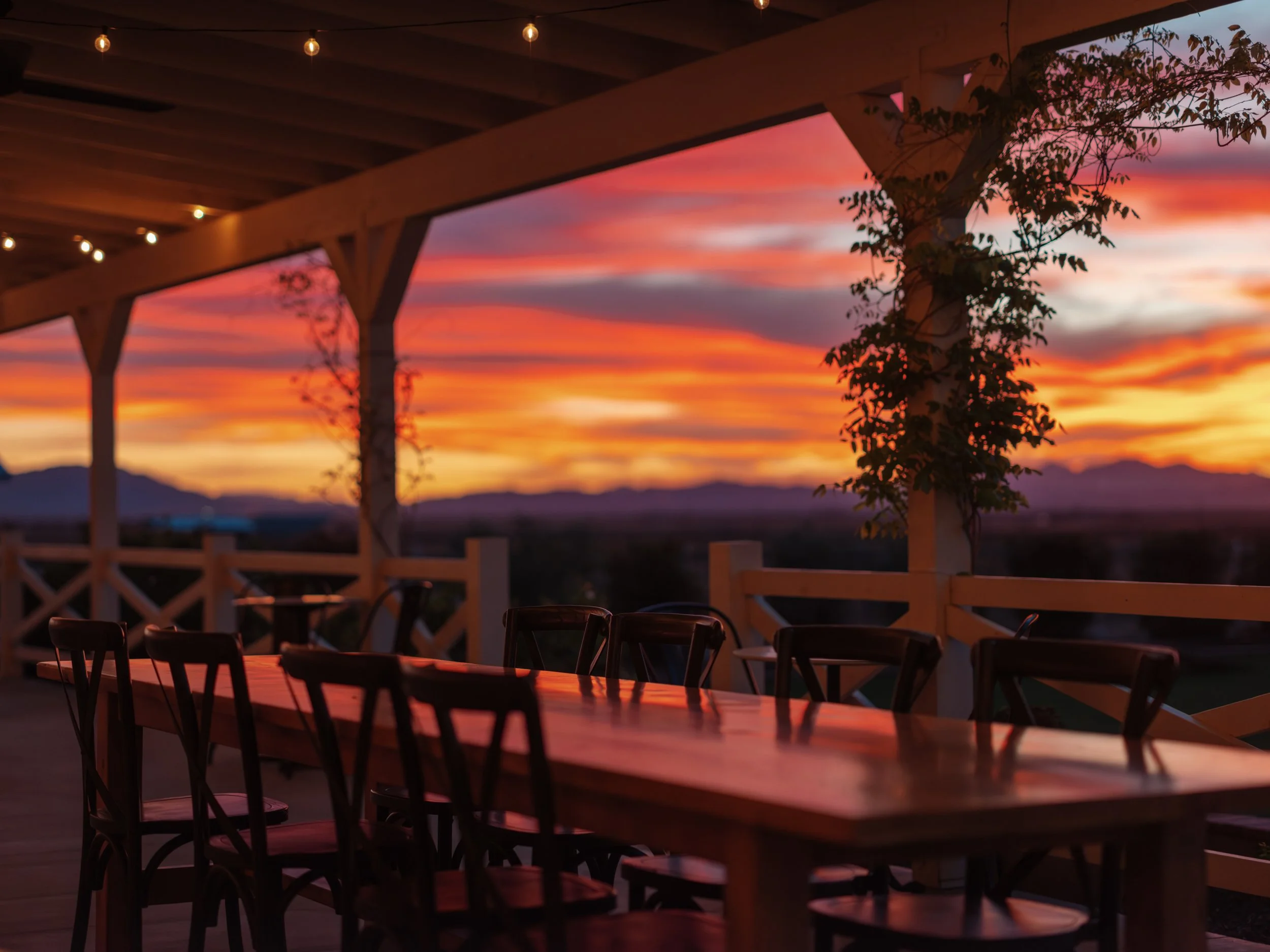 Outdoor patio with a large wooden table and chairs, overlooking a colorful sunset sky with pink, orange, and purple hues, and string lights hanging above.