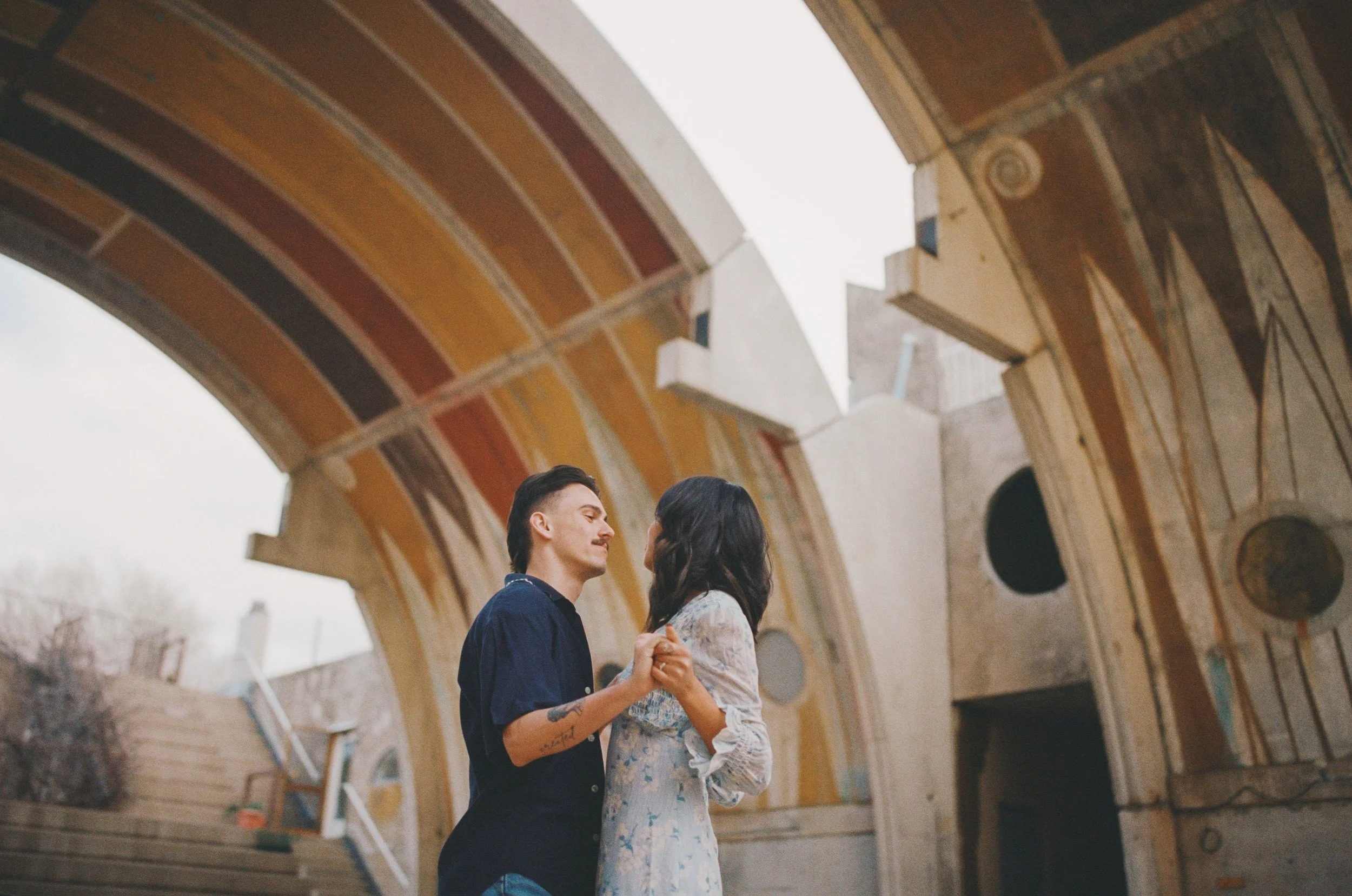 A couple holding hands and looking into each other's eyes under a large, colorful archway with ornate designs.
