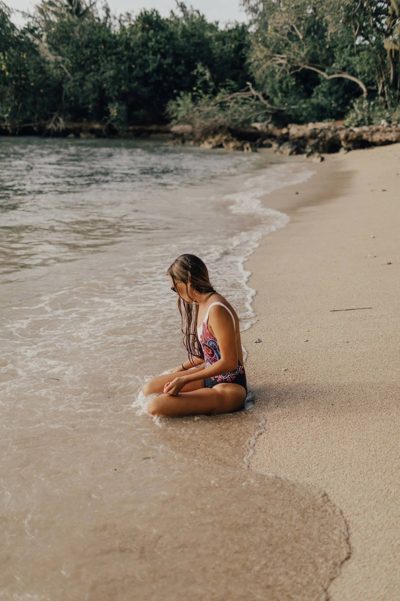 A girl with long hair sitting on the sandy beach near the water's edge, with trees in the background.