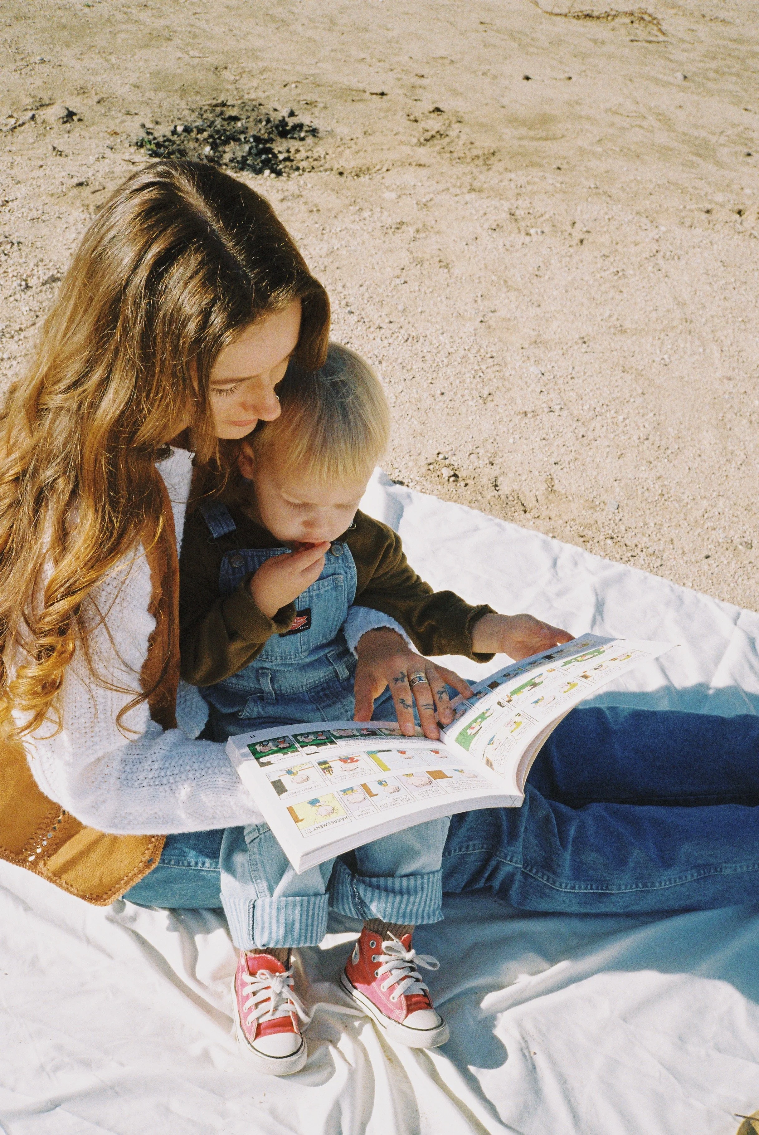 A woman and a young boy sitting on a white blanket on the sand, reading a colorful comic book together.