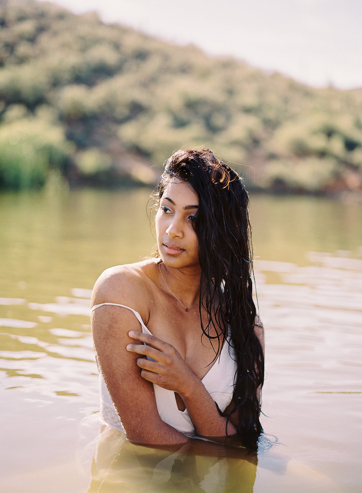 A woman with long dark wet hair standing in a body of water, wearing a white top, with her arm crossed over her chest, outdoors with a blurred natural landscape background.