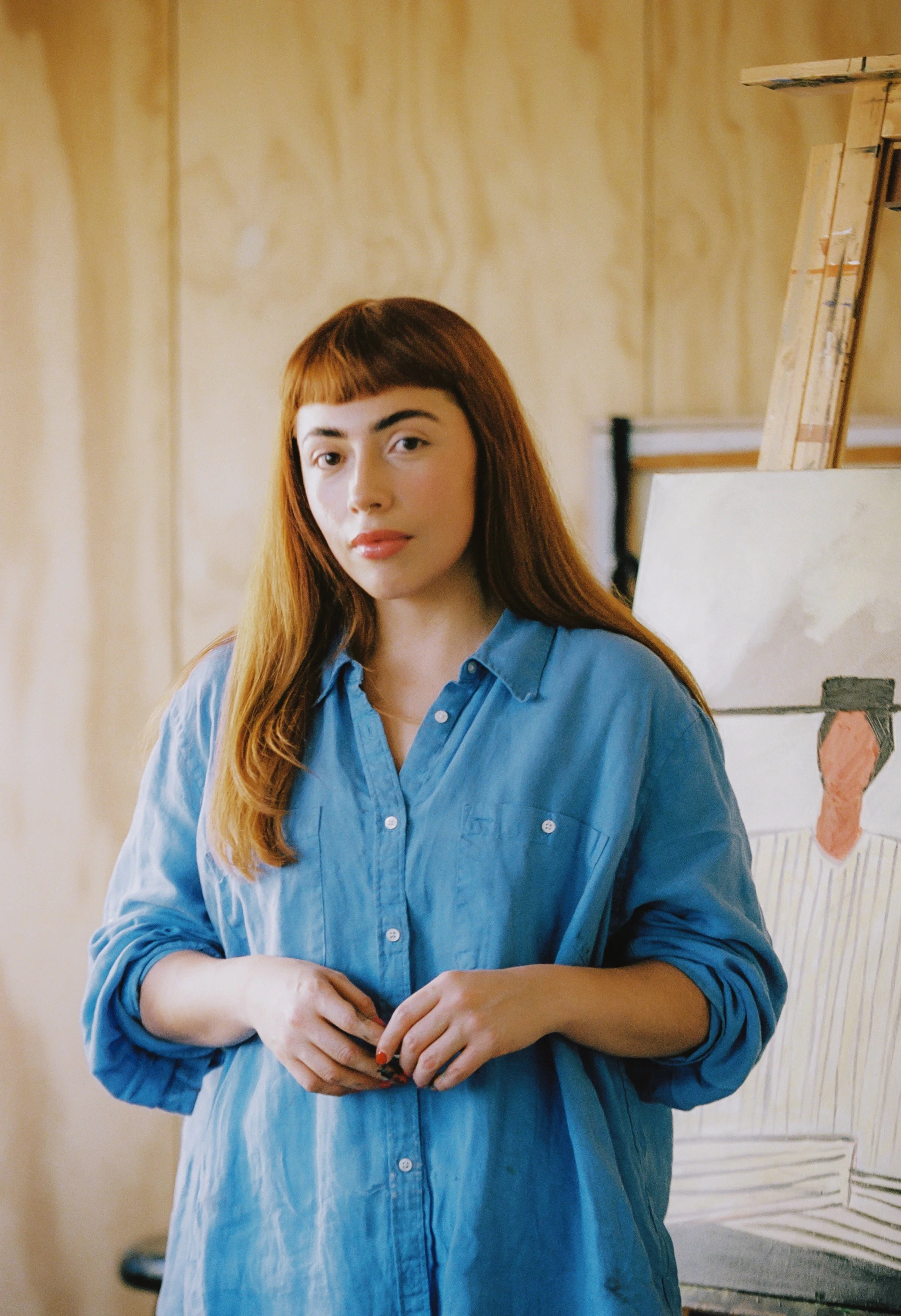 A young woman with long reddish hair and bangs, wearing a blue button-up shirt, standing indoors in front of a plain wall and an art easel with a drawing of an ice cream cone.
