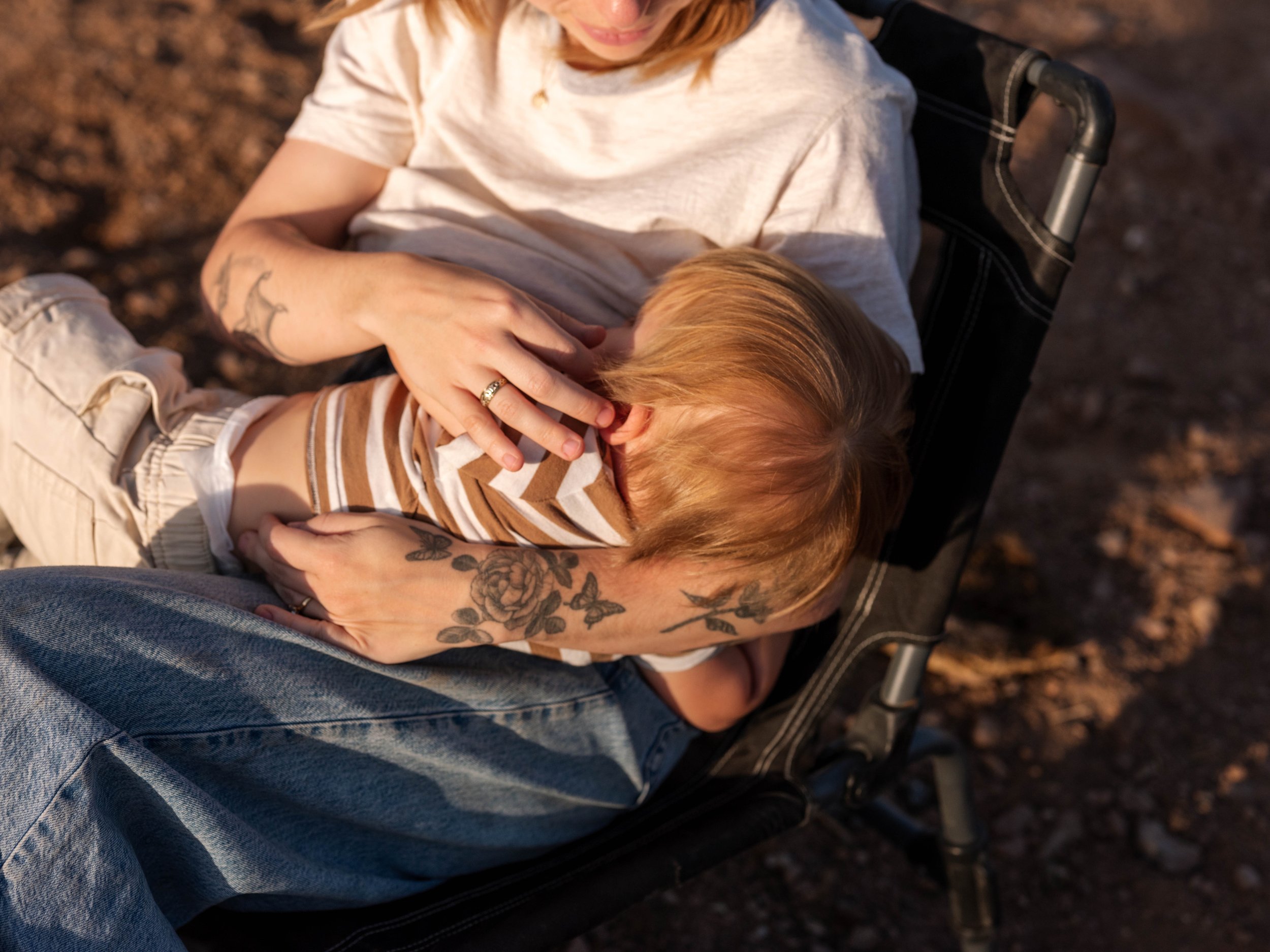 A woman with tattoos and a ring on her finger sits in a wheelchair, holding and cuddling a young child with red hair, wearing a striped shirt, on her lap outdoors with rocky ground.
