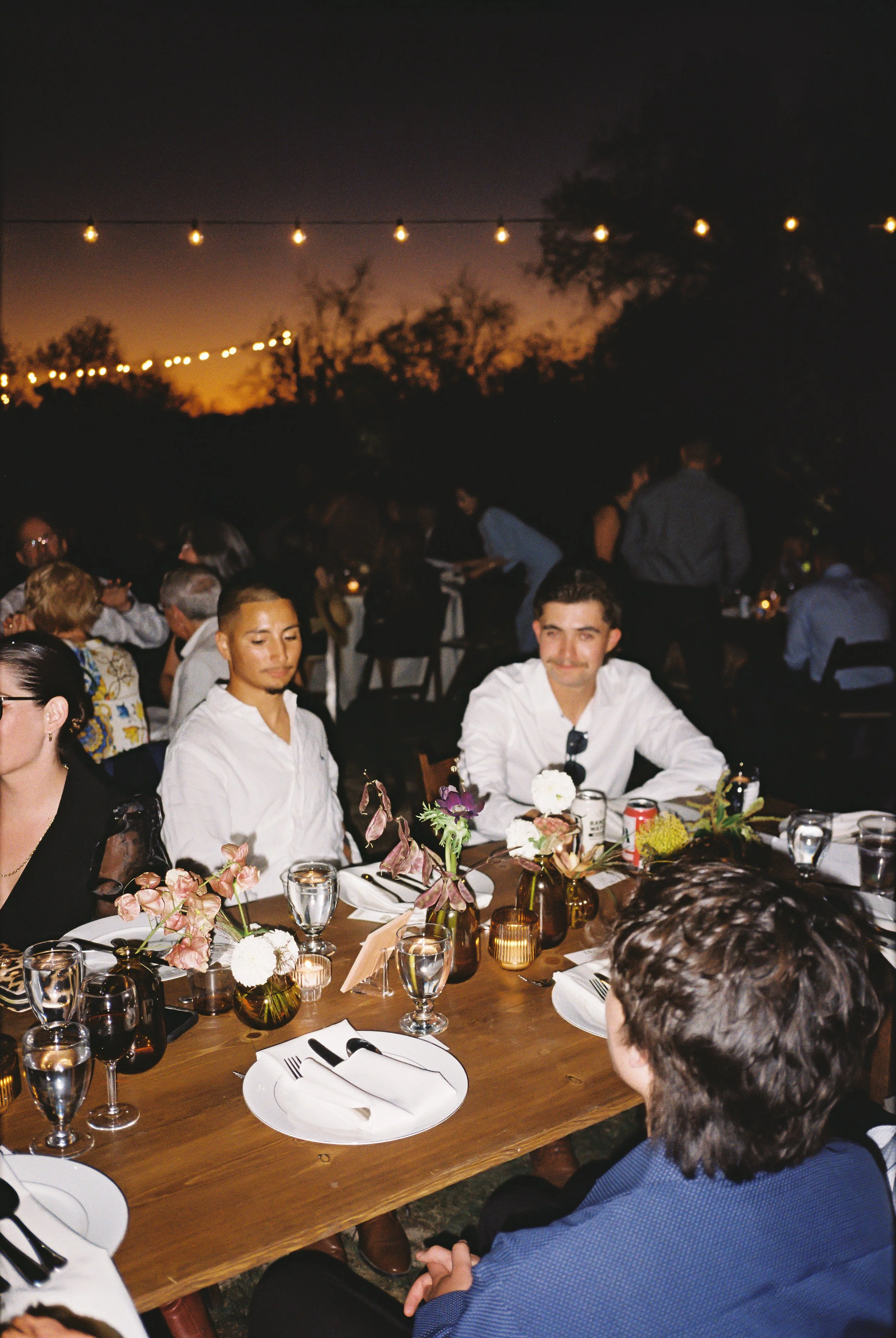People dining at a sunset outdoor dinner party with string lights overhead, decorated table with flowers, glasses, and plates.