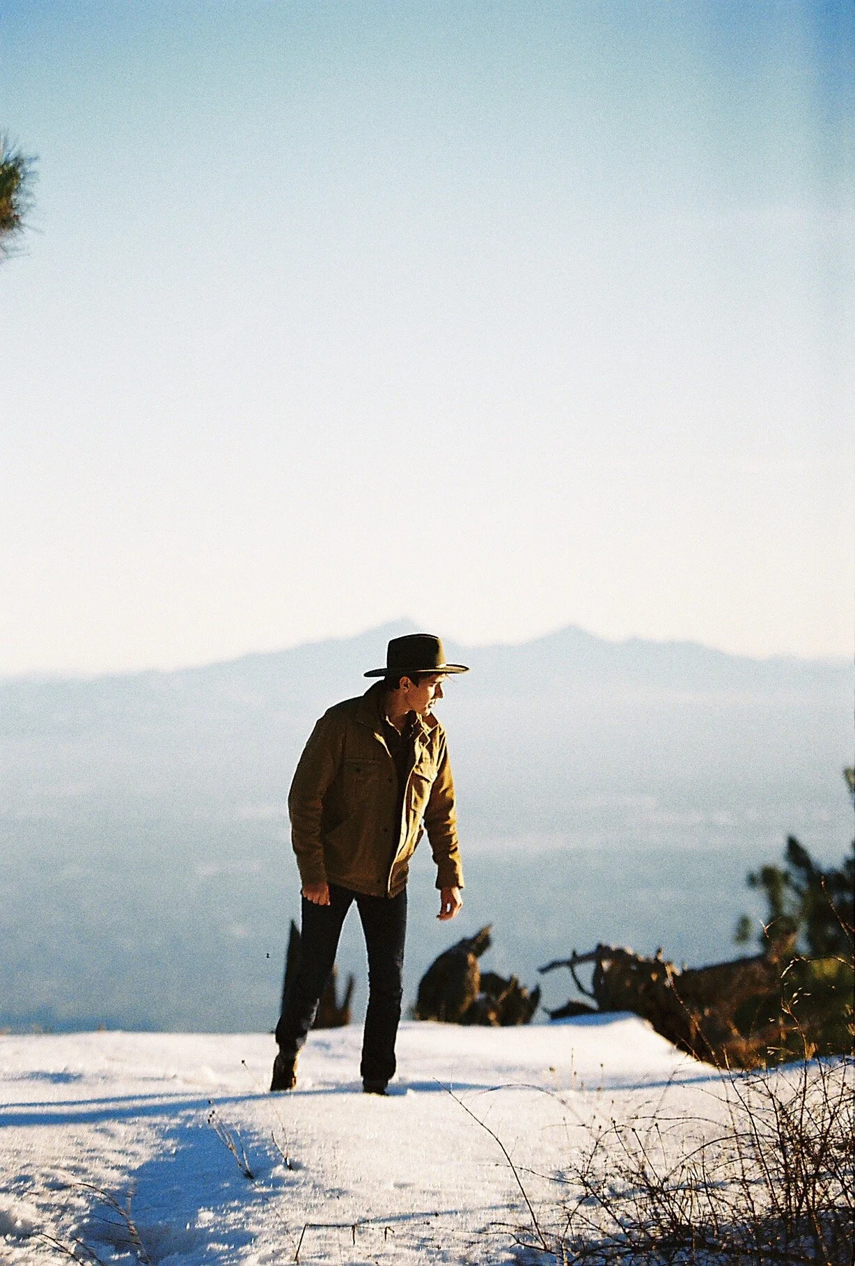 A person wearing a brown jacket and black hat walking on snow-covered ground in a mountainous landscape.
