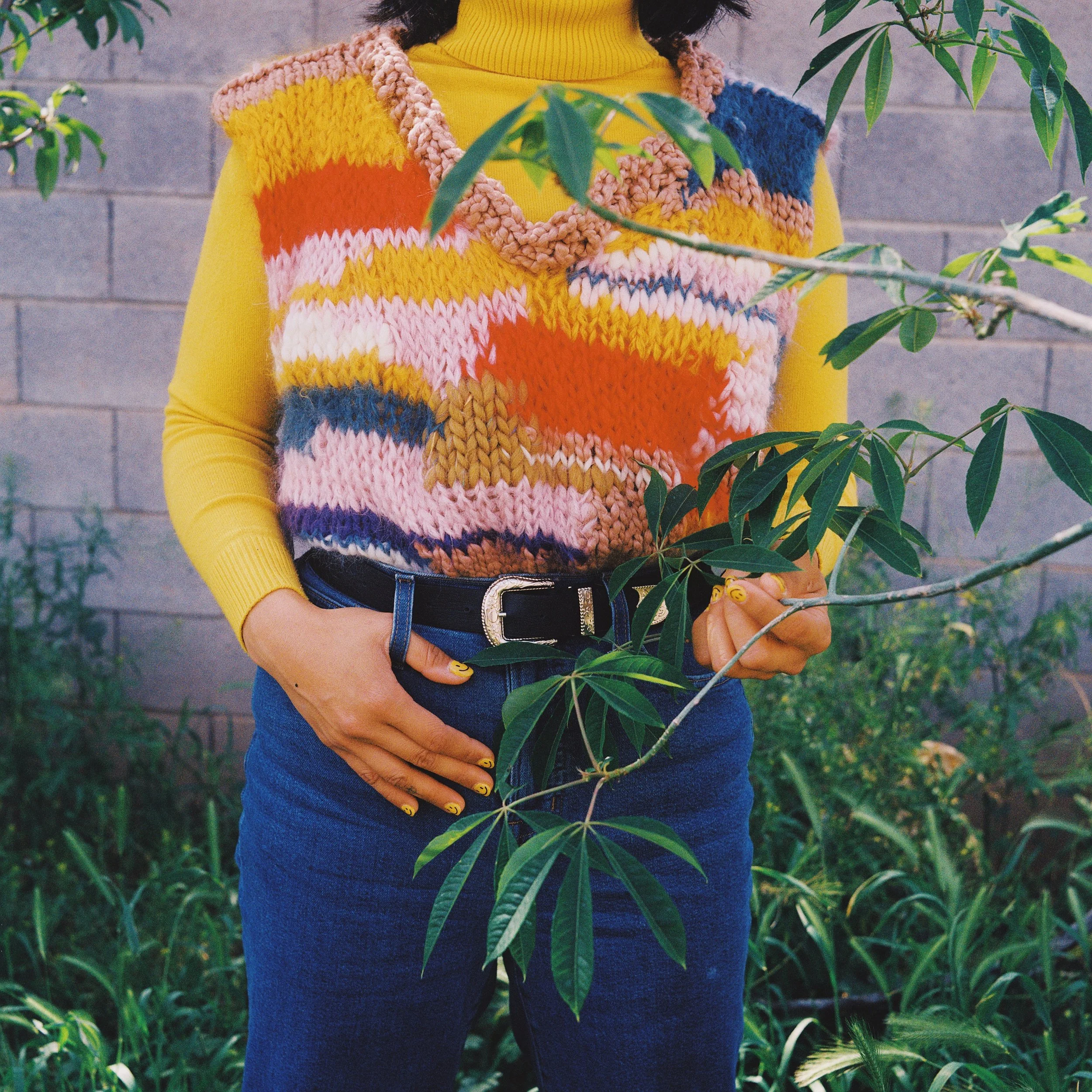 Person wearing a yellow long-sleeve shirt and a multicolored knitted vest, standing outdoors near green plants and a brick wall, holding a branch with leaves.