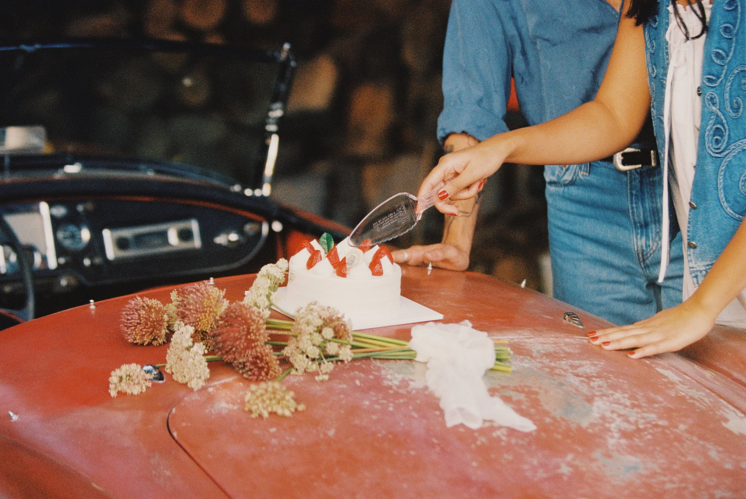 Person decorating a birthday cake with strawberries, surrounded by flowers on a red car hood.