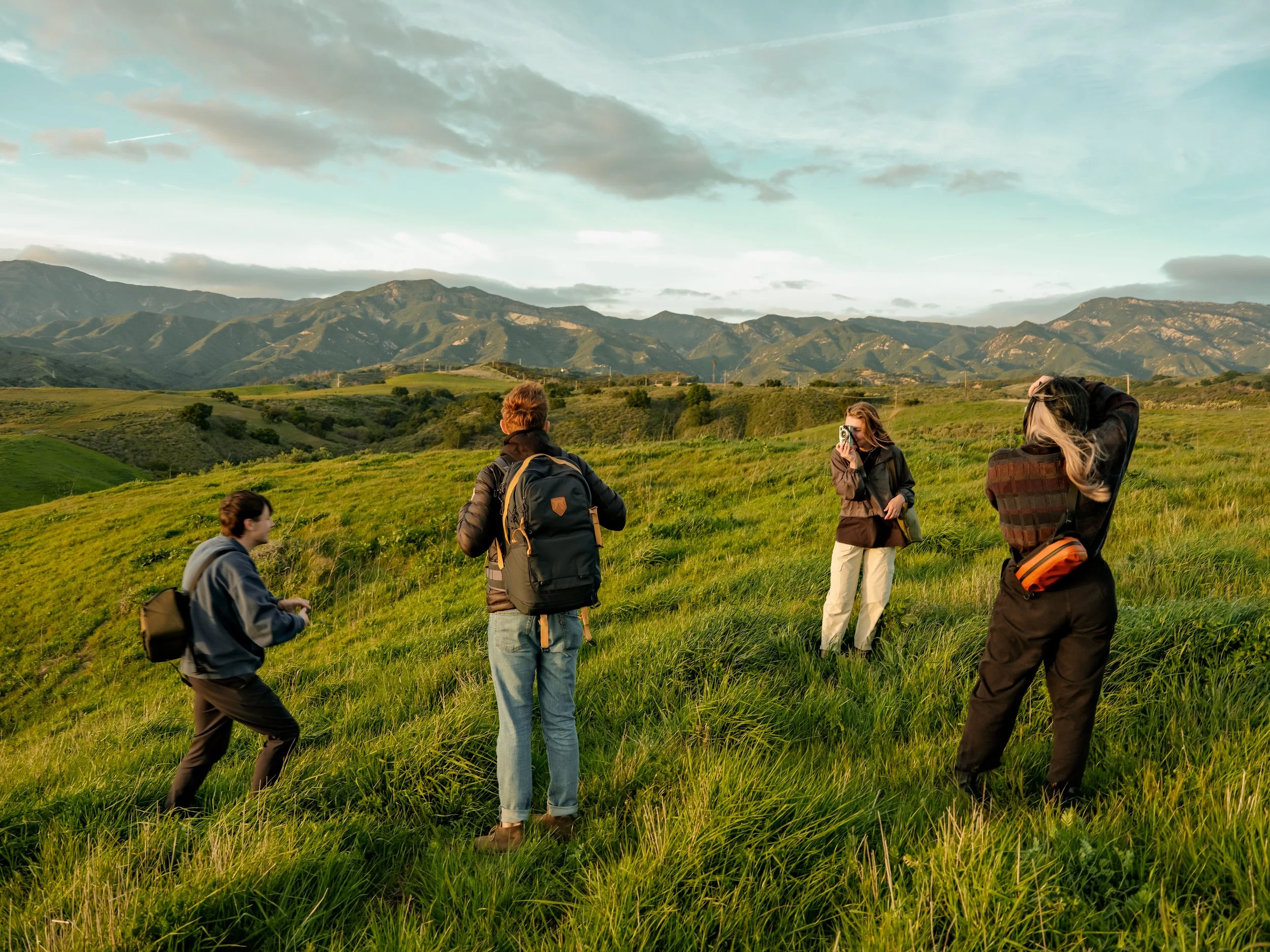 A group of five hikers in outdoor gear standing on green grass in a mountainous landscape during daytime, with mountains and a sky with clouds in the background.