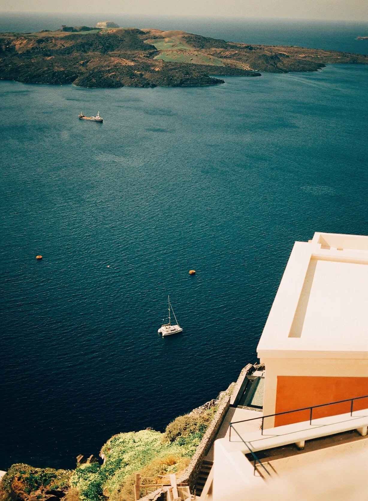 A view of a body of water with a sailboat, two small boats, and a hillside in the background. A white building with a terrace is visible in the foreground.