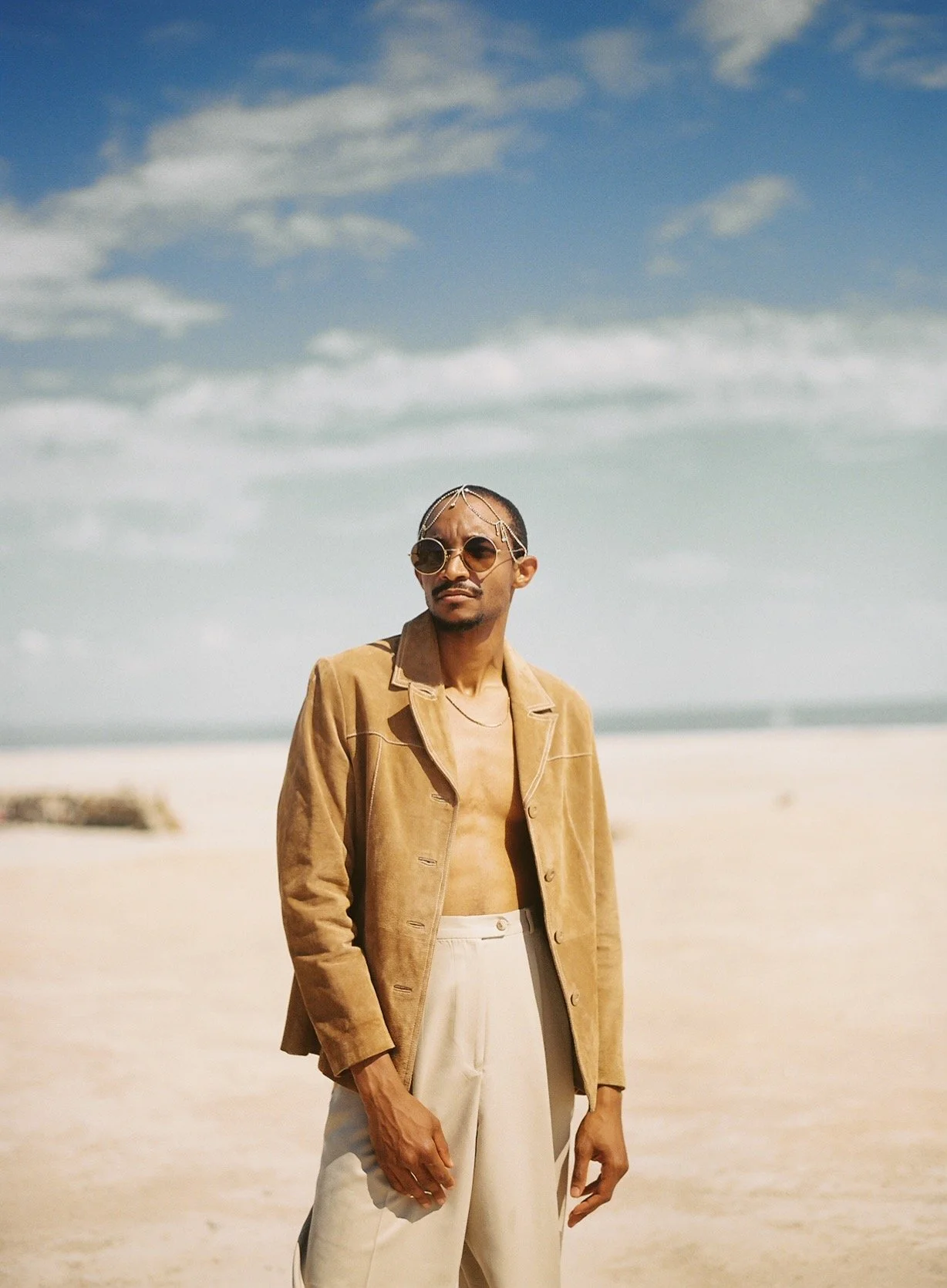 A man standing on a sandy beach with a cloudy sky and ocean in the background. He is wearing a tan jacket, beige pants, sunglasses, and has chains in his hair.