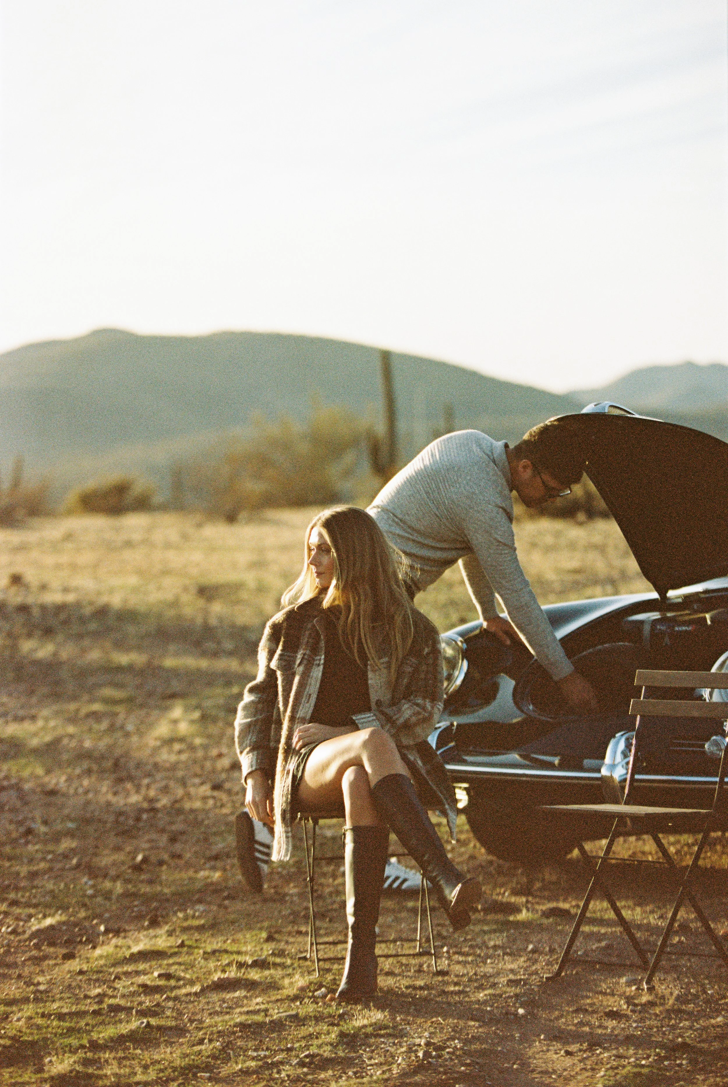 A woman and a man outdoors near a car with its hood open; woman sitting on a folding chair, woman wearing a plaid jacket, black top, and tall boots, man wearing glasses and a light sweater fixing the car.