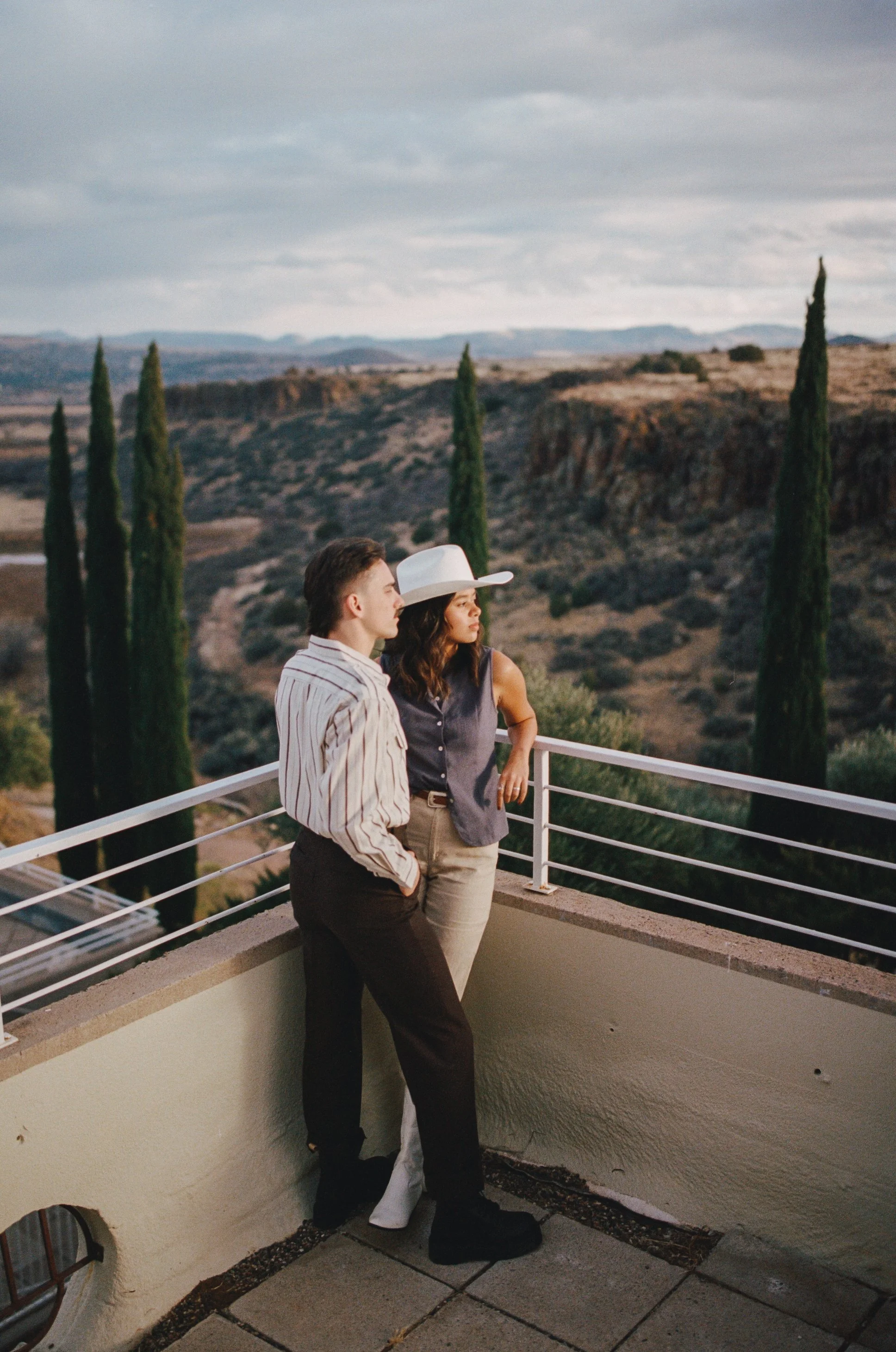 A man and woman stand on a balcony overlooking a scenic landscape of rolling hills and tall cypress trees, with cloudy skies above. The woman is wearing a wide-brimmed white hat, sleeveless top, and beige pants, while the man is dressed in a striped 
