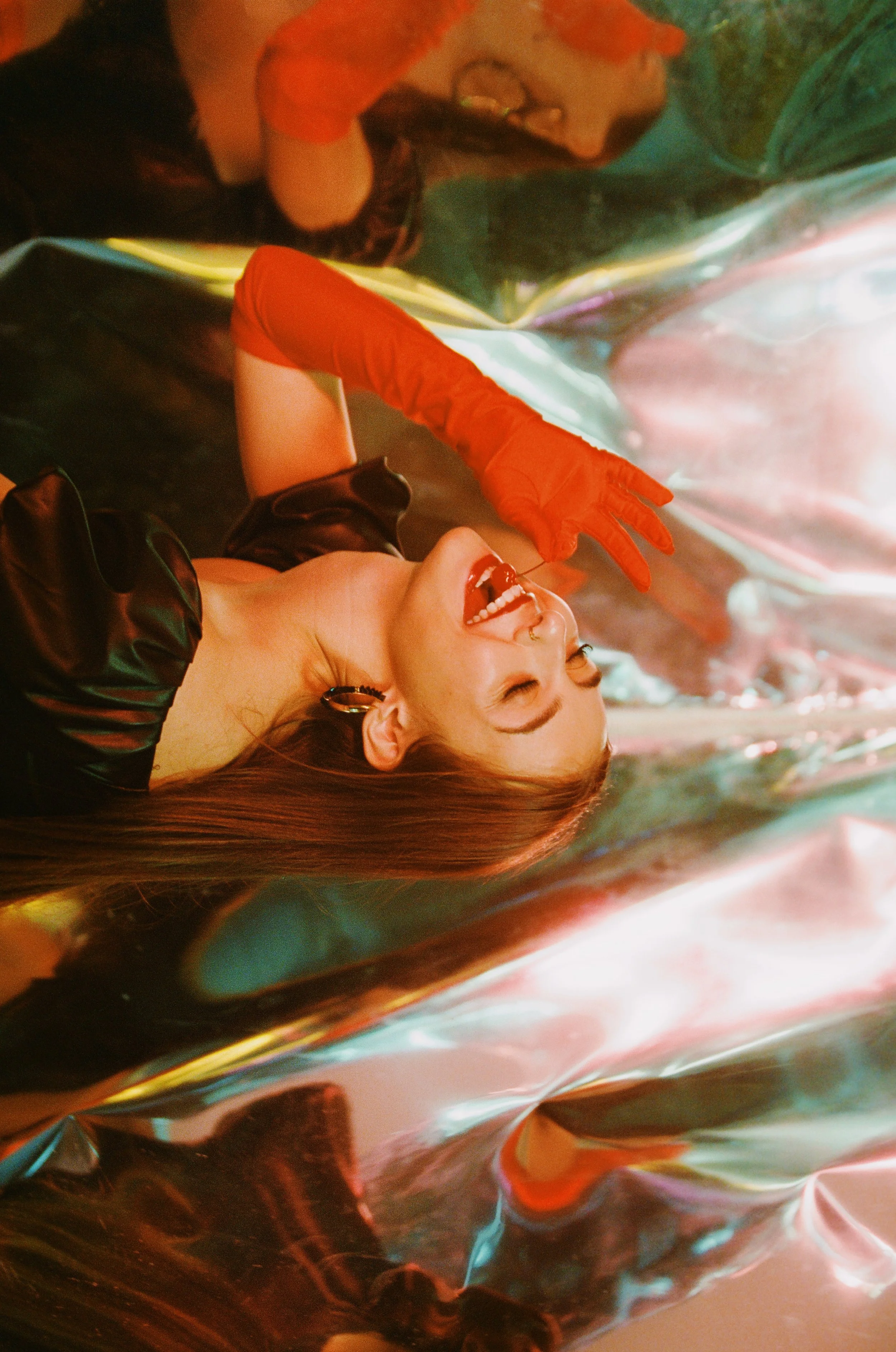 Woman laughing with long red hair, wearing black leather top and red gloves, lying on a shiny, colorful metallic surface.