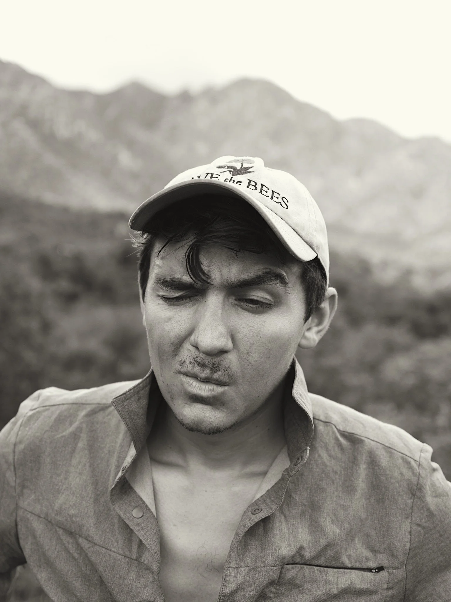 Black and white photo of a young man winking, wearing a cap that says 'Life the Bees' and a button-up shirt, with mountains in the background.