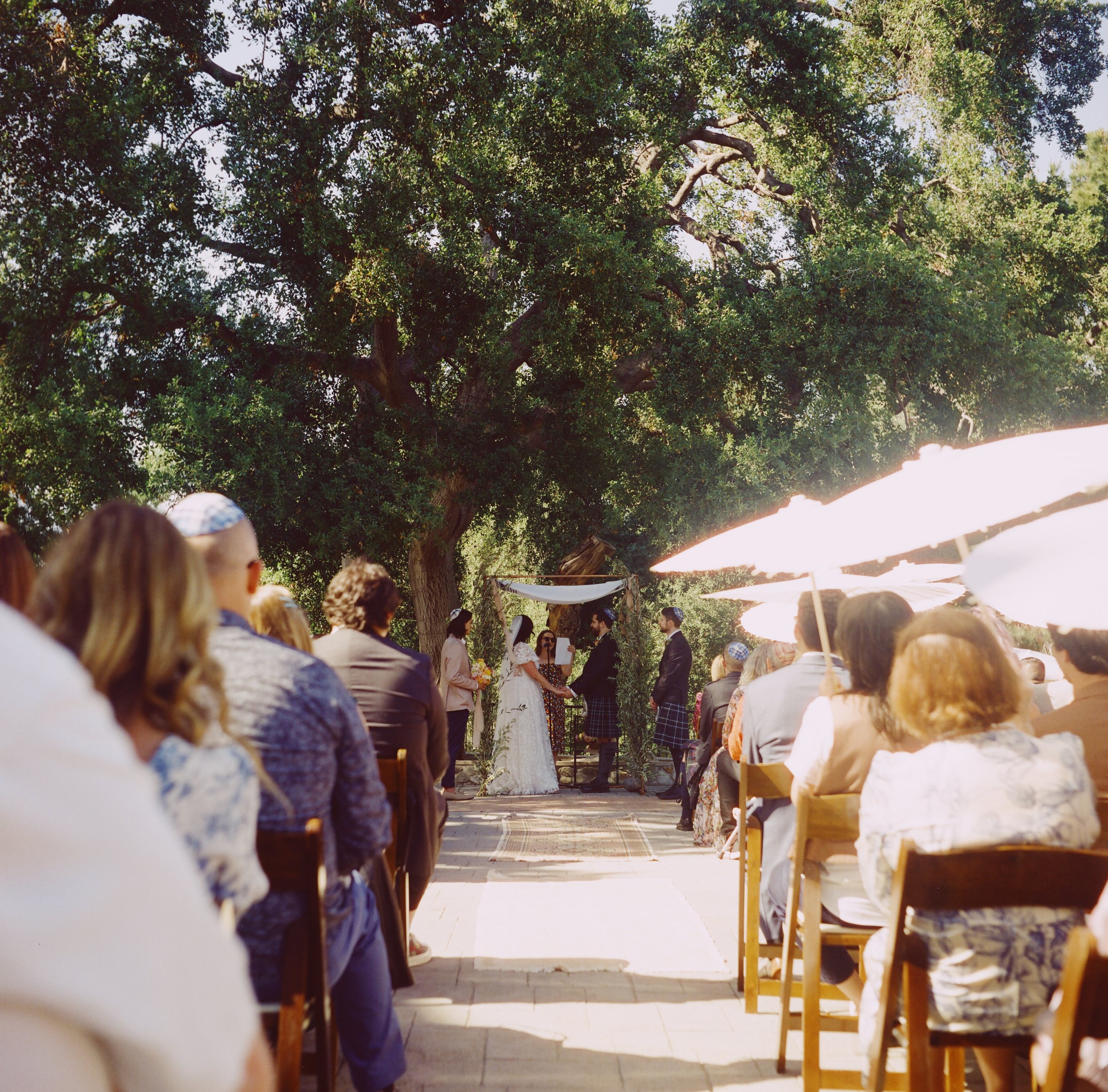 Outdoor wedding ceremony with bride and groom exchanging vows under a canopy, surrounded by guests seated on chairs, with large white umbrellas providing shade, and tall trees in the background.