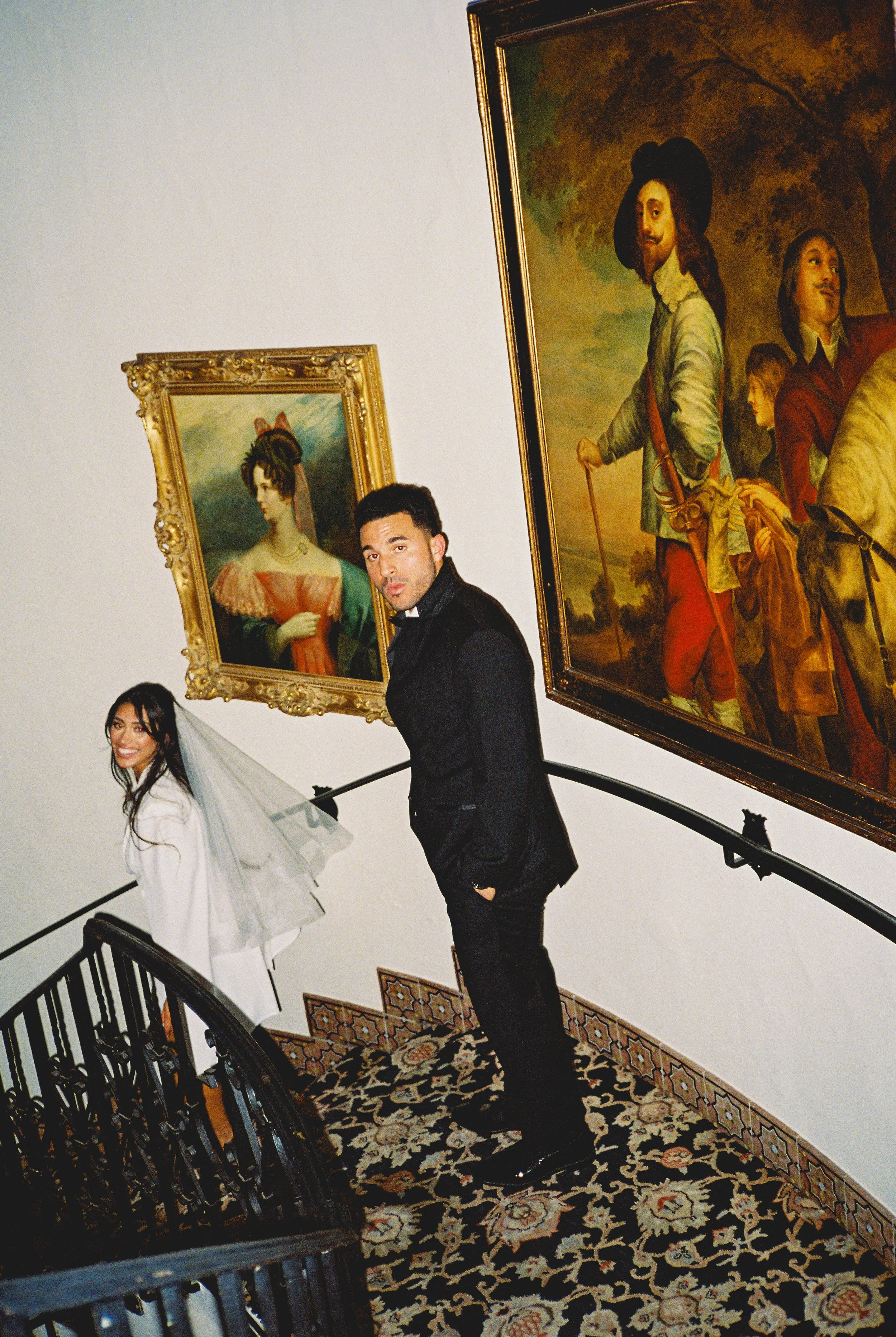 A man and woman standing on a staircase in an art gallery, surrounded by framed paintings of historical figures and portraits.