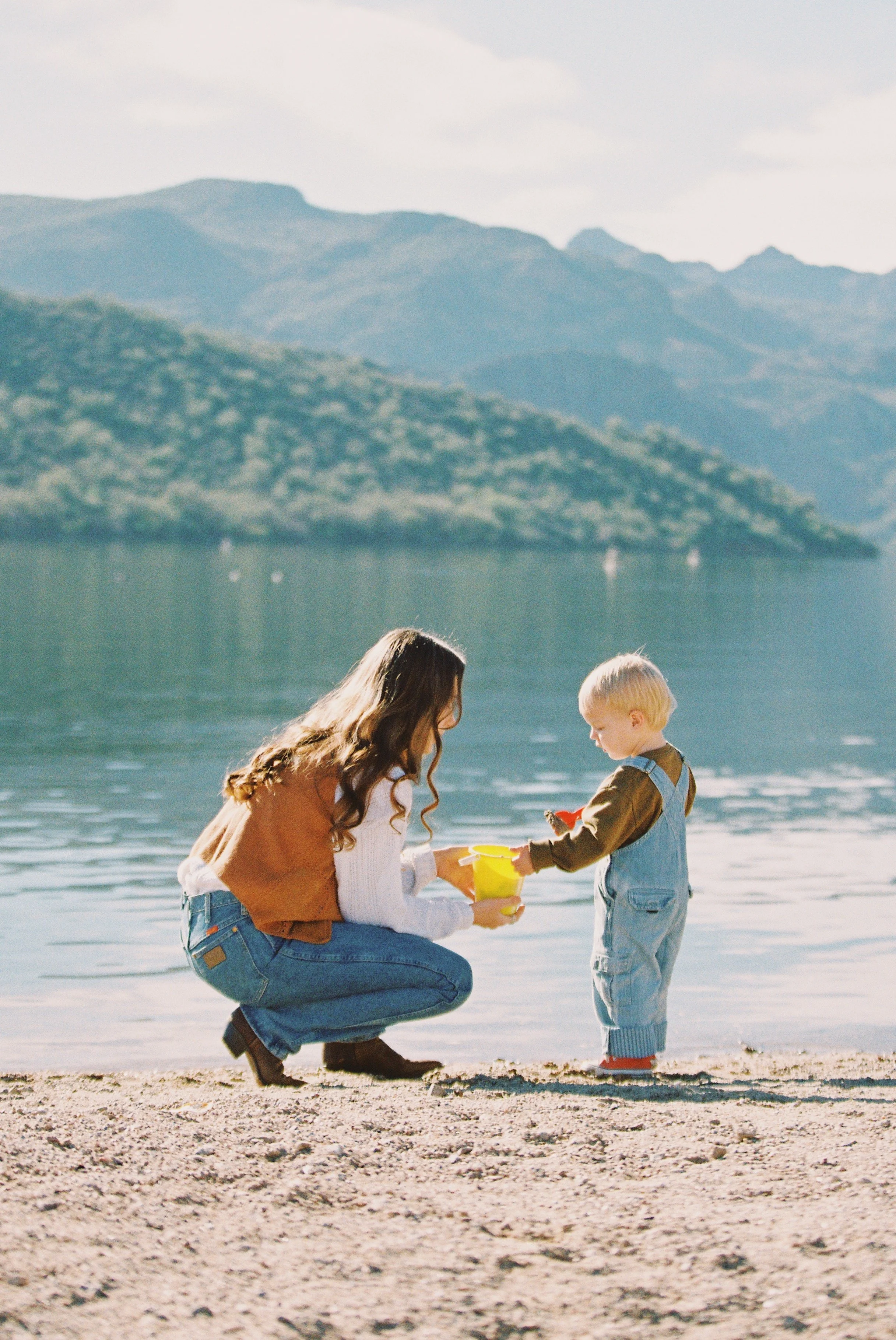 A woman crouching on a sandy beach handing a yellow bucket and a toy to a young boy by a lake with mountains in the background.