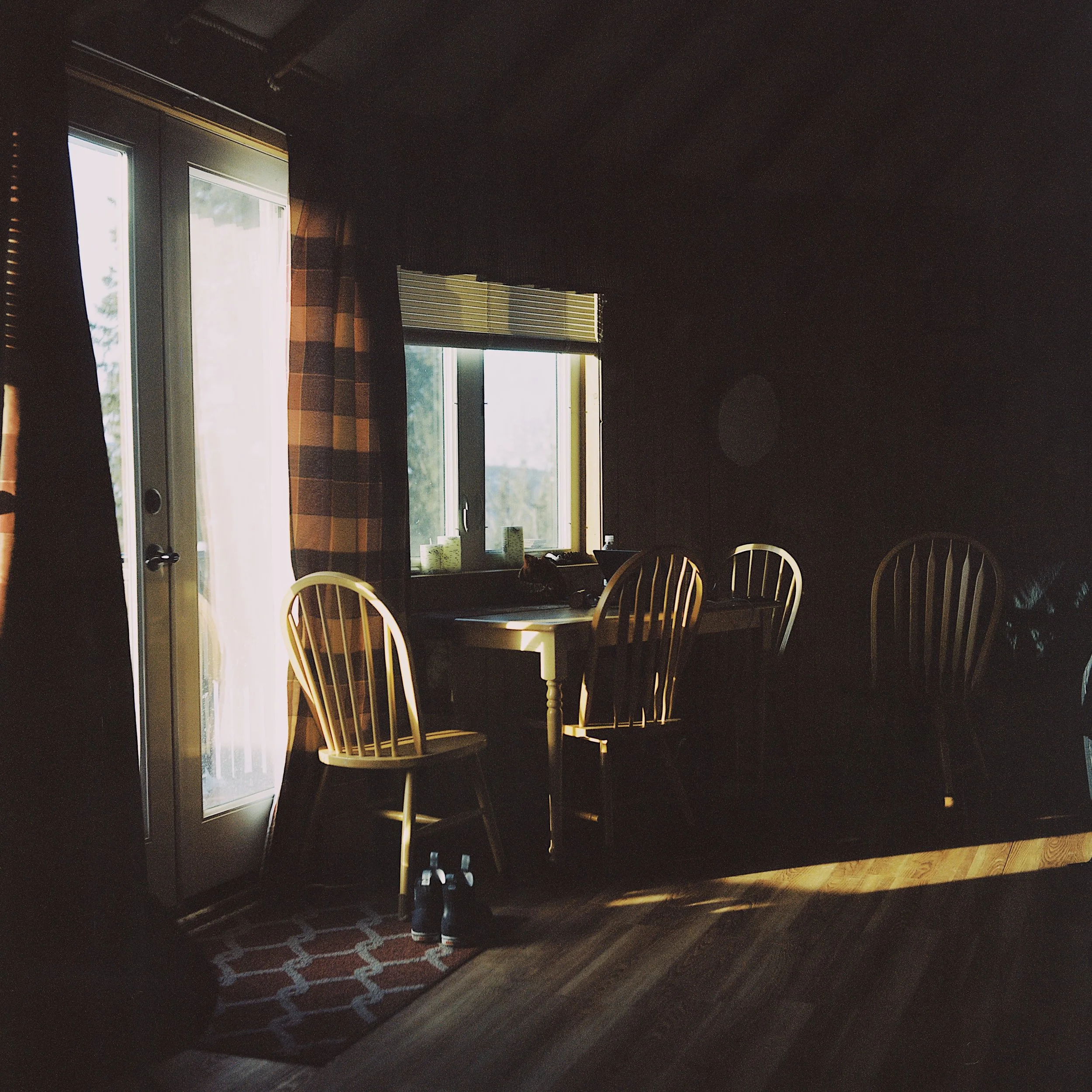 A dark interior room with sunlight coming through a glass door and window, illuminating a wooden table with four chairs, a pair of shoes on the floor, and a patterned rug.