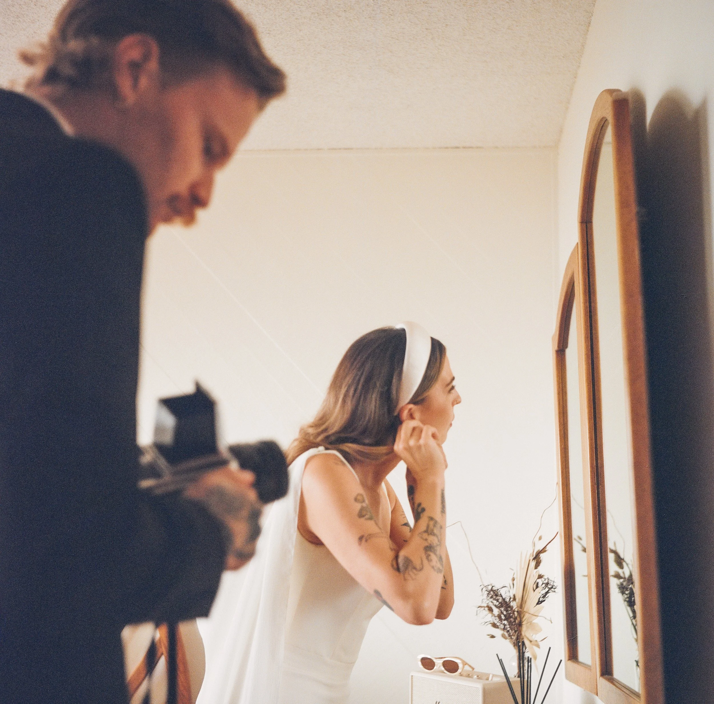 A woman with tattoos on her arm looking into a mirror, smiling, while a man in the foreground, slightly out of focus, appears to be recording or taking a photo with a camera in a room decorated with a vase of dried flowers and sunglasses on a small t